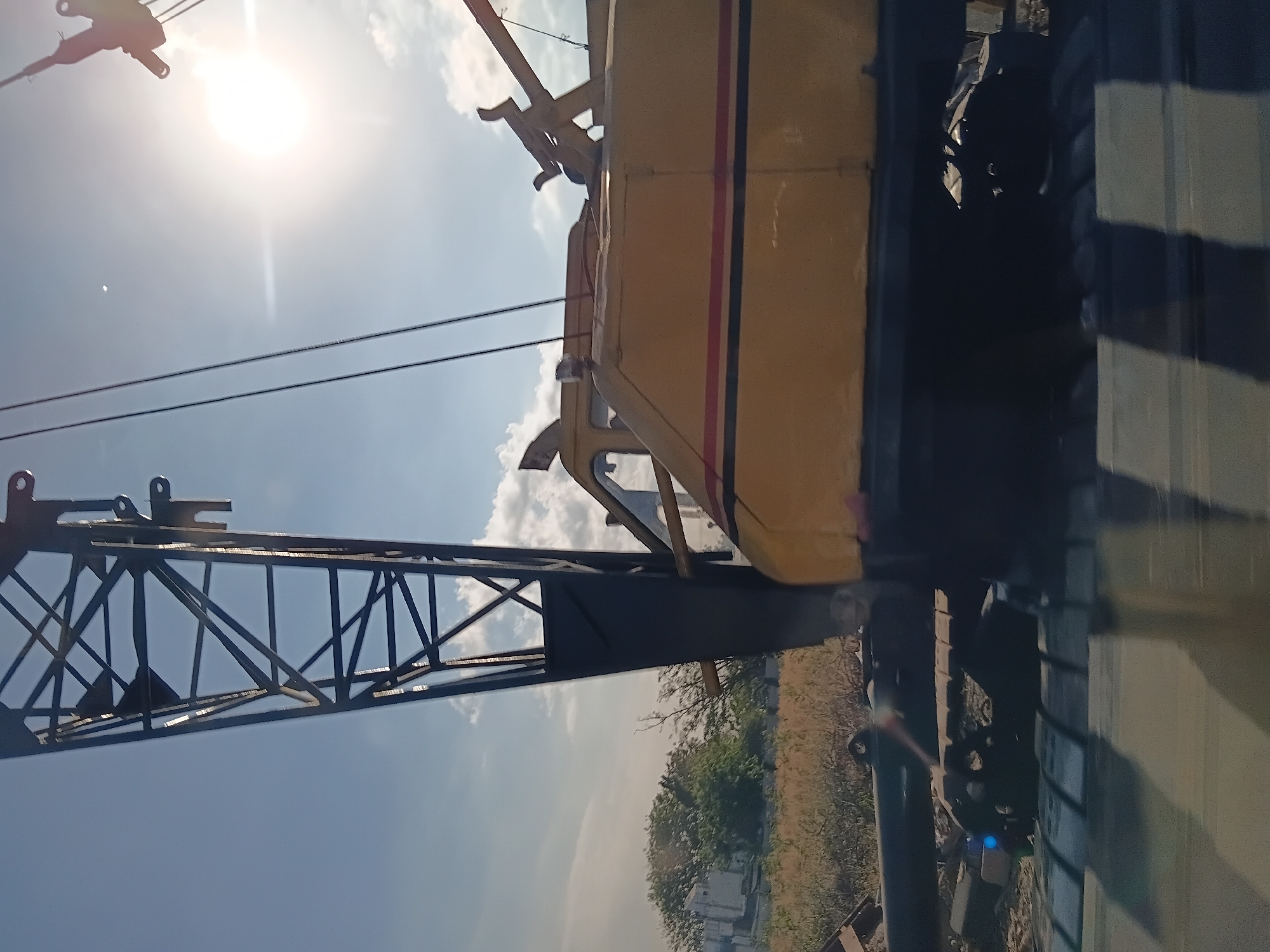 The image shows a large yellow construction crane, partially obscured by the bright sun and sky. The crane's boom is visible in the foreground, extending upwards. The body of the crane, with its distinctive red and black stripe, is also visible. In the background, there are trees and what appears to be a building. The scene is outdoors, under a clear blue sky with some clouds. The image seems to have been taken in Surabaya, Indonesia, based on the context provided.
