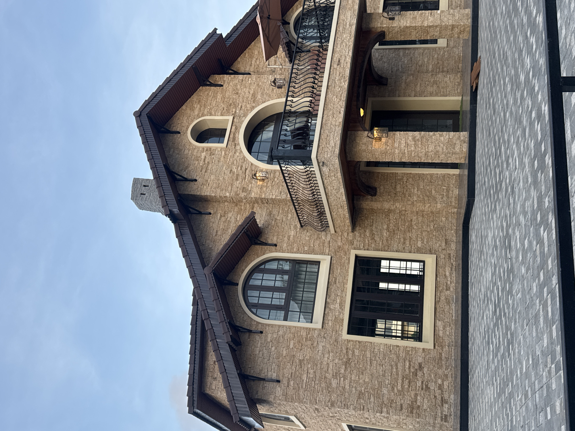 The image displays a two-story residence situated in Xuân Trường - Đà Lạt, Vietnam, under a clear, pale blue sky. The house features a facade constructed from light brown, rough-hewn stone or stone veneer. Its dark brown tiled roof has multiple gables with wide overhangs supported by decorative dark brackets. A stone chimney rises from the left side of the roof.
The house incorporates several window styles, including large arched windows with dark frames and grids on the upper left and above the main balcony. A rectangular window with dark frames is present on the lower left. A prominent second-story balcony extends from the front, featuring an ornate black wrought iron railing and two wall-mounted lanterns. An open brown patio umbrella is visible on the balcony. Below the balcony, the main entrance is dark-framed, flanked by stone pillars, and sheltered by a wooden soffit, also with two wall-mounted lanterns.
The ground in the foreground is paved with interlocking light and dark grey paver blocks arranged in a geometric pattern. The overall scene depicts a well-constructed and maintained property.