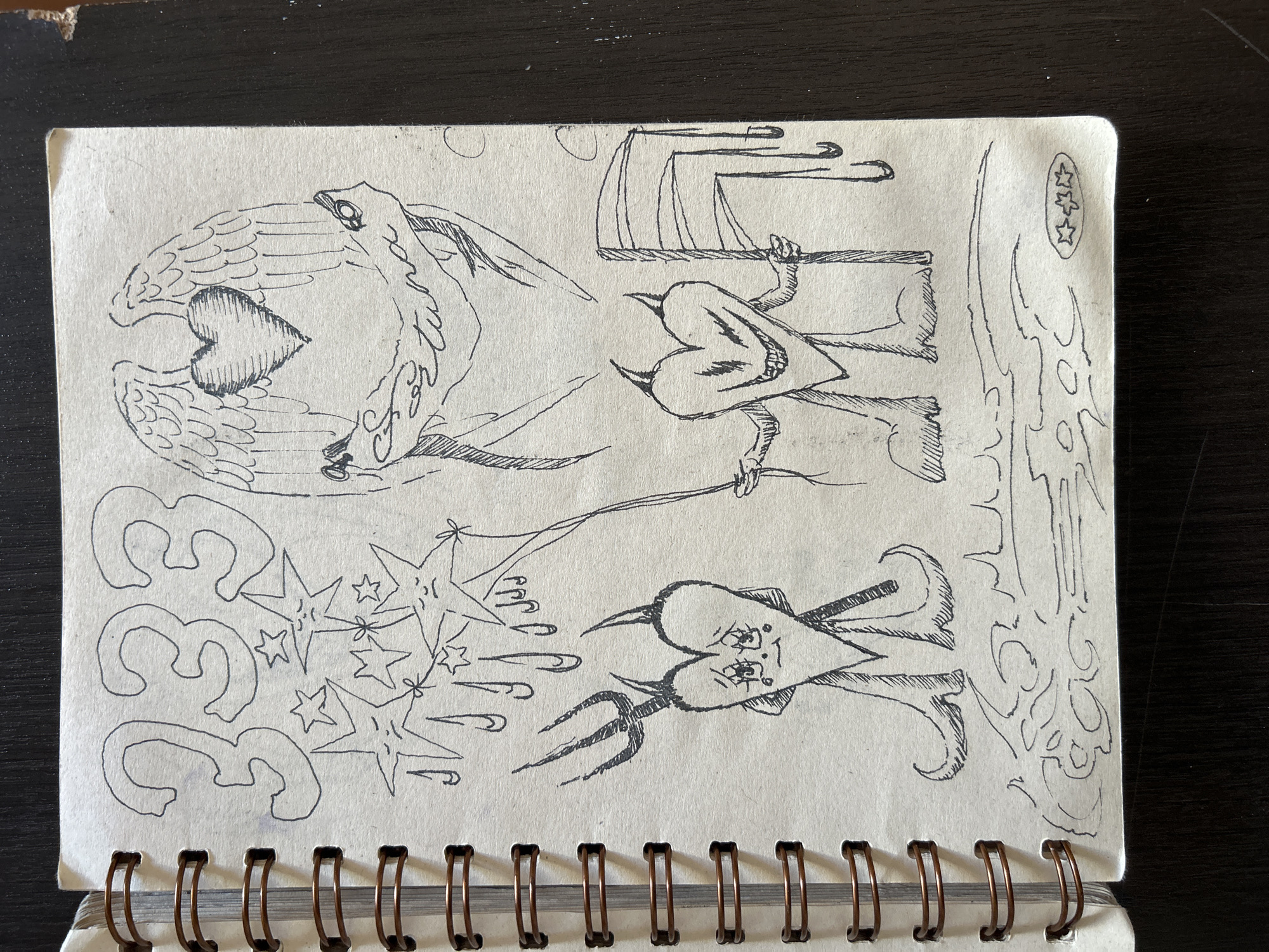 An open, spiral-bound sketchbook page features a collection of whimsical black ink drawings against a light, off-white paper background. The page is resting on a dark, textured surface, with the metal spiral binding visible along the bottom edge.
The page is divided into four main visual sections, each containing distinct illustrations. In the top left, two stylized bird-like figures with prominent wings face each other, cradling a shaded heart between their beaks. Their bodies extend into elegant, flowing lines.
To the right, a heart-shaped figure with limbs and simple facial features (eyes and a beak-like nose) holds a large scythe. It stands beside an abstract structure composed of vertical and horizontal lines, resembling a minimalist altar or chair. In the top right corner of the page, three small, outlined stars are stacked vertically within an oval border.
In the bottom left quadrant, the numbers "333" are boldly outlined and stacked vertically. Adjacent to these numbers, a cluster of stars of varying sizes appears to be in motion, with lines emanating from them. One of the larger stars contains a small, indistinct figure.
The bottom right section depicts another heart-shaped figure, this one resembling a devil, complete with small horns, a pointed tail, and a trident held in one hand. Its face, drawn on the heart, has defined eyes and a mouth. To its right, an abstract, swirling pattern that resembles flames or clouds fills the corner.
All drawings are rendered in a clean line-art style with occasional cross-hatching for shading, creating a collection of symbolic and fantastical imagery.