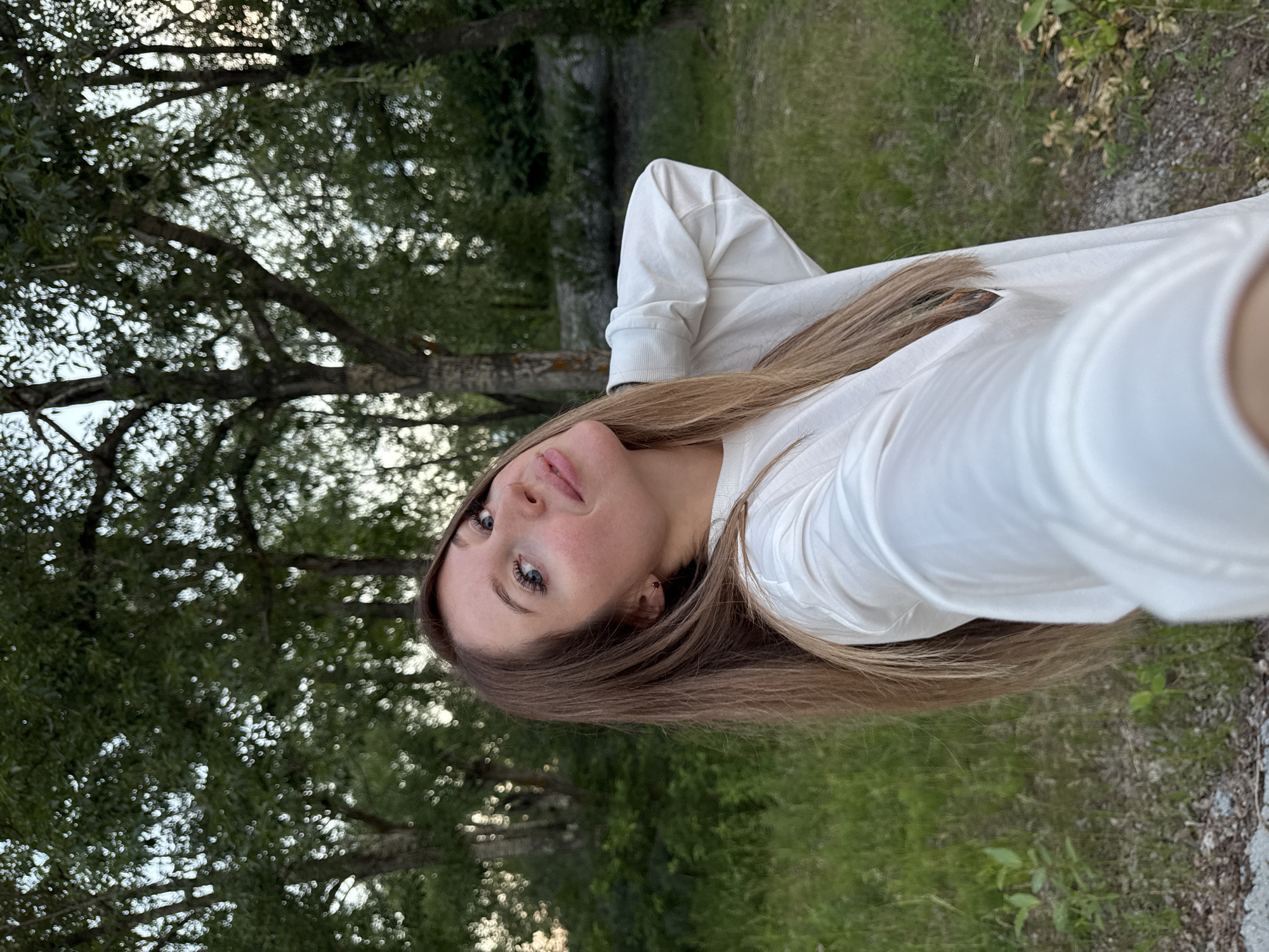 A young woman with long, straight brown hair is taking a selfie outdoors. She is wearing a white long-sleeved shirt. The background consists of trees with green leaves and tall grass, suggesting a natural setting within or near the city of Chaikovsky, Russia, possibly in a park or wooded area. The lighting indicates it is daytime, likely late afternoon or early evening due to the soft light.