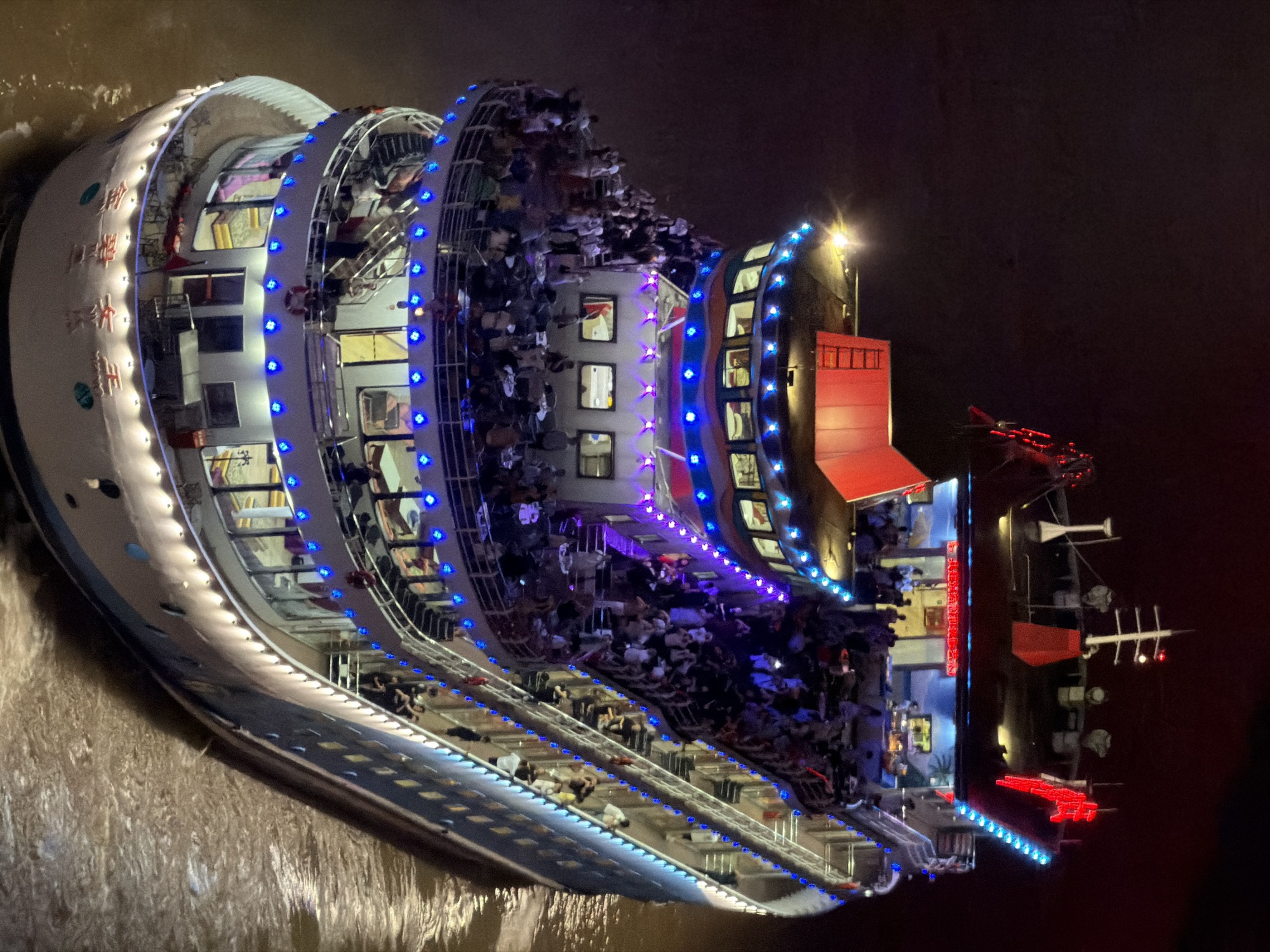 The image shows a multi-decked passenger ship illuminated by numerous blue and red lights at night. The ship is crowded with people on its open decks and visible through the windows of its enclosed levels. The ship appears to be navigating on a body of water, with brown, rippling water visible around its hull, suggesting it is a river. The ship's exterior is decorated with bands of blue and red lights, and red accents are visible on its upper structures. Chinese characters are visible on the ship's hull, possibly indicating its name or destination. The overall scene depicts a lively night cruise on a busy waterway.