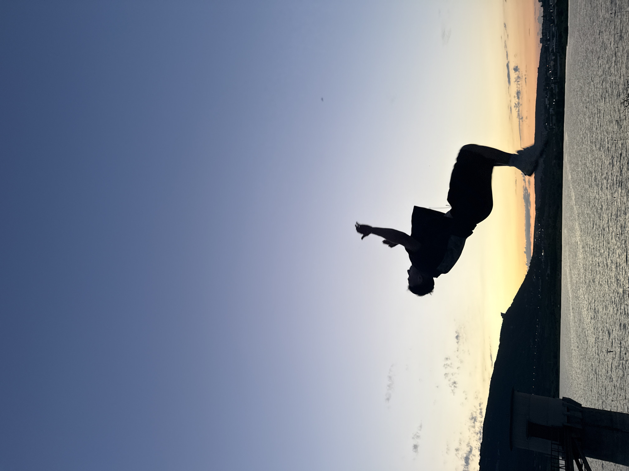 A silhouette of a person performs a backflip against a twilight sky at the Shumen Reservoir in Shumen, Bulgaria. The person, clad in a t-shirt and shorts, is suspended mid-air, arms outstretched. In the background, a large hill or mountain range forms a dark silhouette against the fading light. A tower-like structure, possibly part of a waterworks or observation point, stands to the left of the frame, its concrete base and upper section clearly visible. The reservoir's surface reflects the warm hues of the sunset. The sky transitions from a deep blue at the top to lighter shades of blue and orange near the horizon.