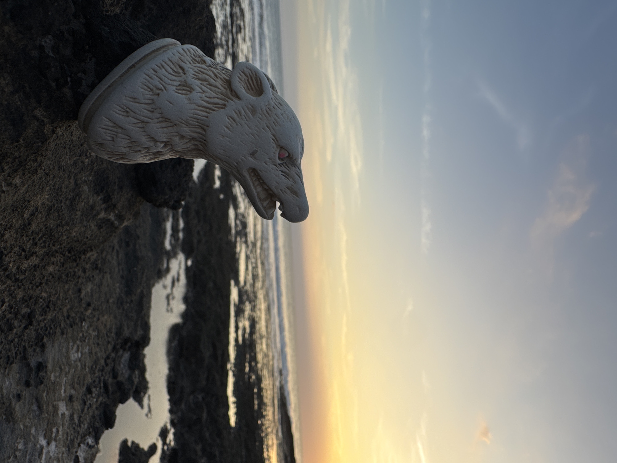 A white wolf head statue sits on a rocky, uneven surface, possibly a coastal outcrop. The statue is positioned on the left side of the frame, facing towards the right, with its snout and teeth visible. The eyes appear to be red. The backdrop consists of an ocean horizon under a sky transitioning from yellow-orange near the water to light blue above. This scene is potentially located on the coast of Kediri, Indonesia. No people are visible in the image.