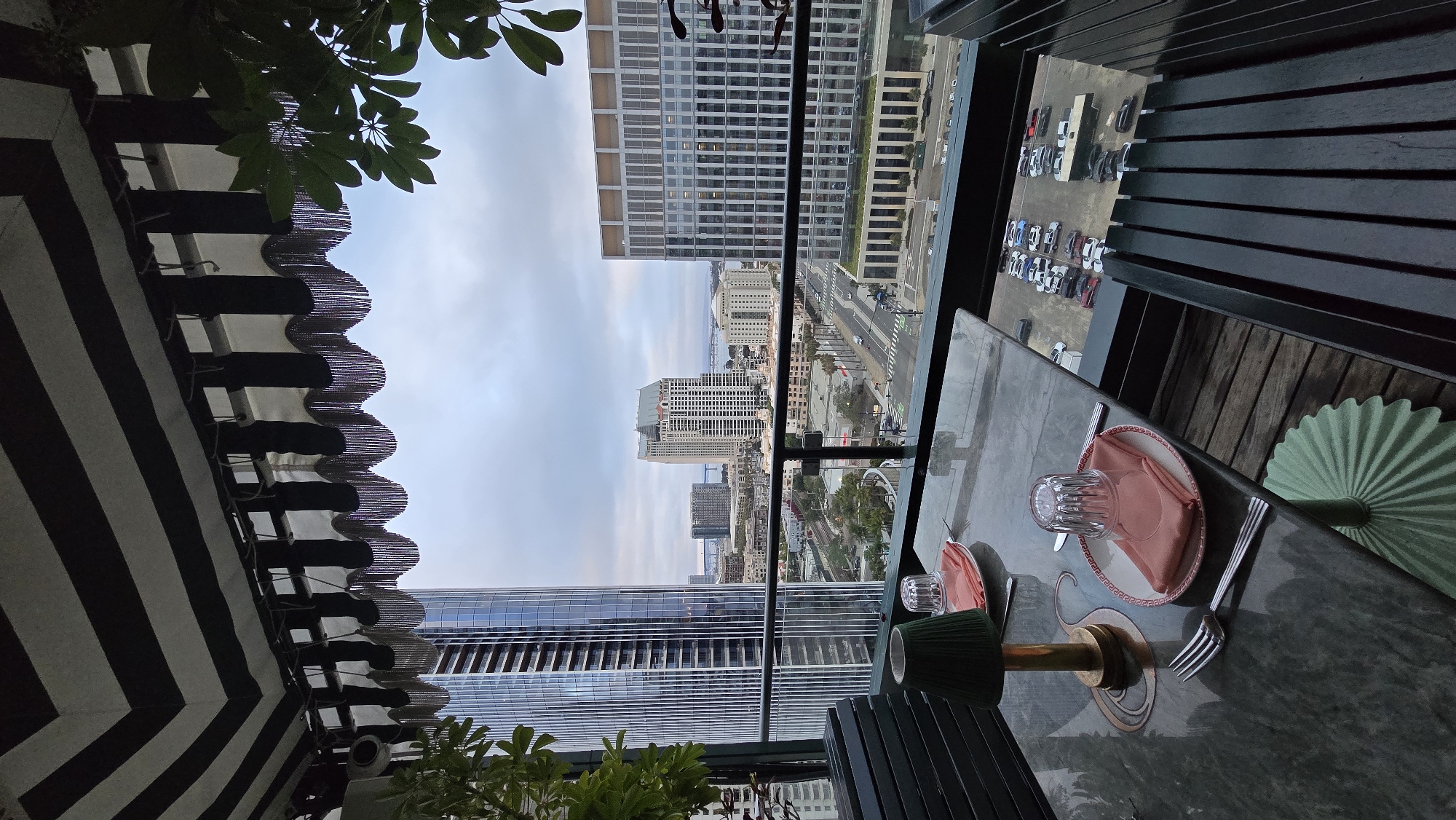 A medium shot captures a table set for dining on a balcony overlooking a cityscape under an overcast sky. The table, with a polished dark grey surface, holds two place settings. Each setting includes a white plate with a red and white patterned rim, a folded peach-colored napkin, a clear glass, and silverware. A small, dark green lampshade with a brass base sits on the table, casting a dim light. In the background, a modern city skyline is visible, featuring tall glass skyscrapers and lower buildings. A street with cars and palm trees runs through the middle ground. The balcony itself is partially covered by a black and white striped awning with decorative fringe. Dark green chairs are positioned around the table. The scene appears to be in downtown San Diego, USA, possibly a rooftop restaurant or bar.