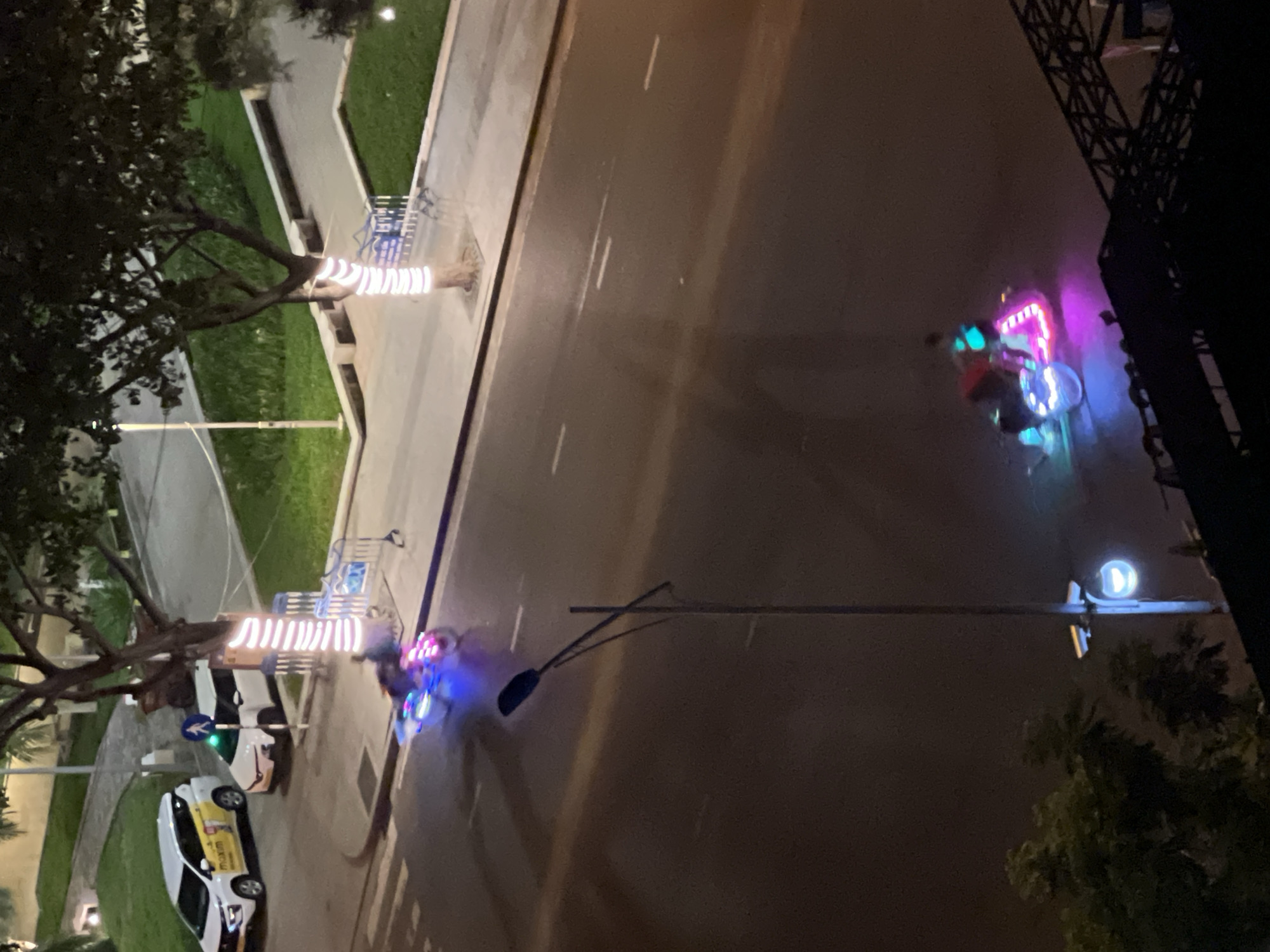 The image shows a street scene at night in Nha Trang, Vietnam. A white car with a yellow stripe and "maxim" branding is parked on the side of the road, next to a white car. A traffic light with a pedestrian symbol is visible. Two motorcycles with colorful LED lights are on the road. One motorcycle is in the foreground and is illuminated with pink, blue, and green lights. Another motorcycle, further back, also has blue and pink lights. A street lamp with a curved fixture and a glowing light is visible on the right side of the frame. Trees with lights wrapped around their trunks are also present. The scene appears to be in an urban area with sidewalks and green spaces.
