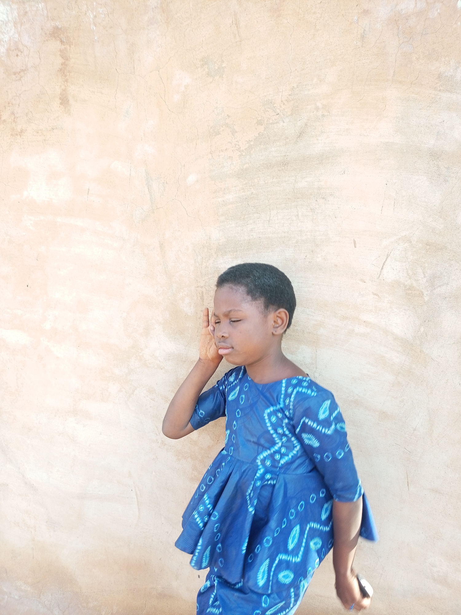 A young Black child, appearing to be around 5-7 years old, stands against a textured, light brown wall. The child is wearing a vibrant blue dress with a white tie-dye pattern. Their left hand is raised to their face, covering their eye, and their eyes are closed. The child is facing to the right of the frame, with their body angled slightly away from the viewer. The background wall shows signs of wear, with cracks and variations in color and texture. The lighting suggests an outdoor setting with sunlight casting a shadow on the child's face. The location is identified as Tempure, Nigeria.