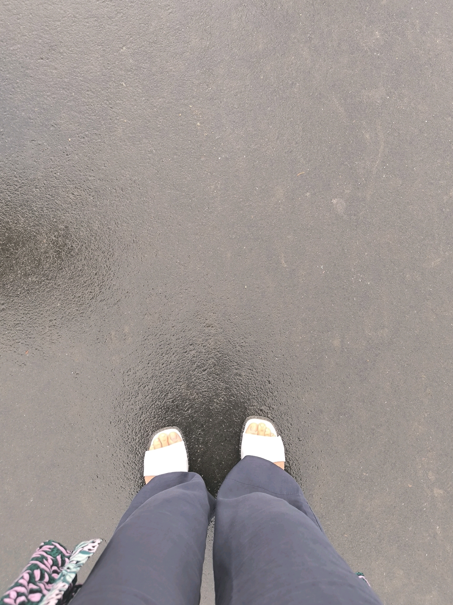 This is a close-up, overhead shot of a person's legs and feet standing on a wet, dark asphalt surface, possibly in Novosibirsk, Russia. The asphalt has a textured, granular appearance with reflections of light, indicating it is damp. The person is wearing wide-leg, dark navy trousers and white slide sandals. A portion of a patterned bag or garment, featuring green, pink, and white colors, is visible on the left side of the frame, near the person's legs. The background is entirely composed of the asphalt surface, with no other elements visible. The image is focused on the lower extremities of the person and the ground beneath them.