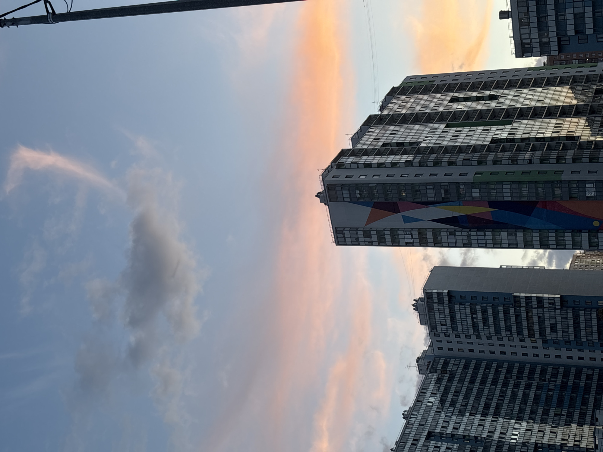 The image presents a modern urban landscape under a colorful sky, likely at sunrise or sunset. The sky, oriented horizontally, transitions from a pale blue to soft pink, orange, and yellow hues across a prominent cloud band that stretches across the mid-ground. Scattered, wispy grey clouds are also visible.
Dominating the right side of the frame are two tall, contemporary buildings, likely apartment complexes, with a third partially visible in the distance. The most prominent building features a dark, multi-windowed facade with balconies and stepped architectural elements. A distinctive, vibrant geometric mural in shades of red, blue, yellow, and green adorns the lower visible section of this building, adding a striking artistic element. The second building, positioned below and slightly behind the first, exhibits a similar modern design with numerous windows. Both structures reflect the soft light and colors of the sky.
In the upper left corner, a dark utility pole or arm with associated wires is partially visible against the light blue sky. The scene is static, devoid of people or discernible activity. The overall impression is one of modern architecture bathed in the warm, serene light of either dawn or dusk. No visible text is present.