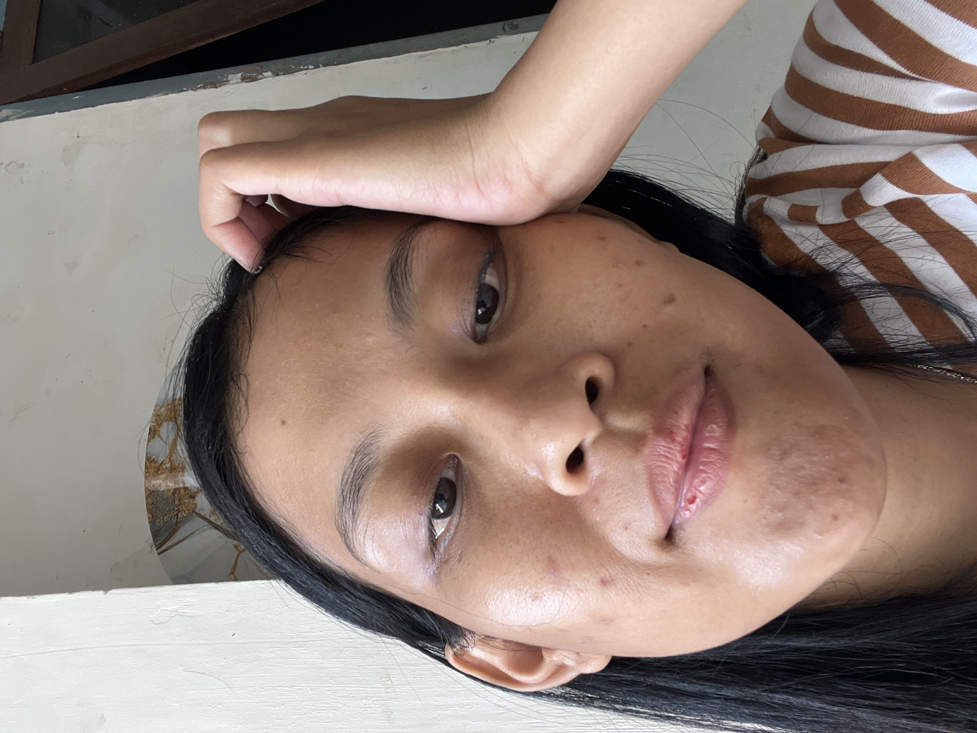 A close-up, eye-level, outdoor, selfie shows a young Indonesian woman with medium-toned skin, long, straight black hair, and brown eyes, resting her head on her right hand. She has light brown eyebrows, and her face exhibits some minor blemishes and acne. She is wearing a brown and white striped t-shirt. The background consists of a white painted wall with some water stains, and a wooden framed window on the right. The scene appears to be in a residential area in Kecamatan Pati, Indonesia.