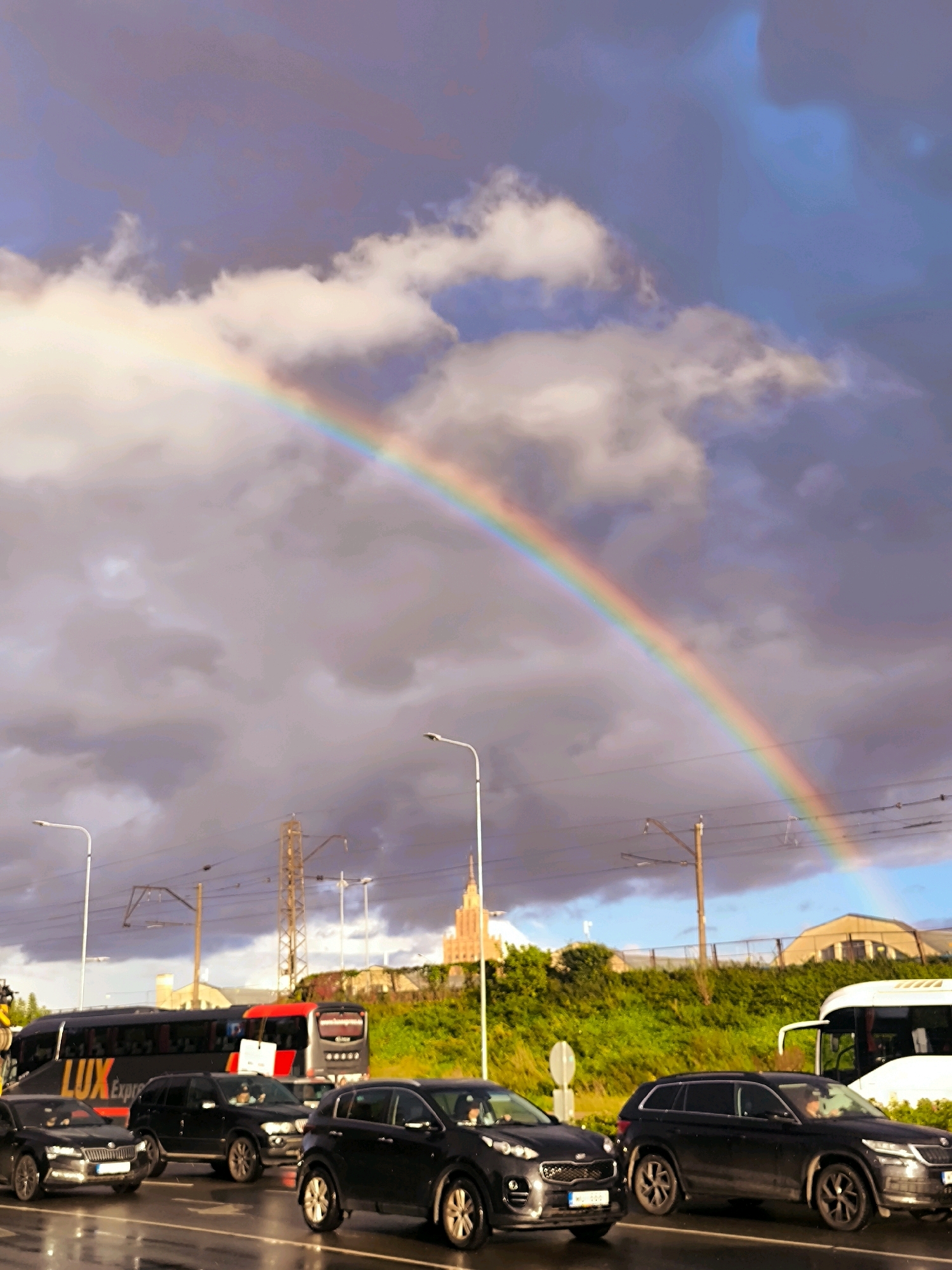 A picturesque rainbow arches across a dramatic sky filled with dark, brooding clouds and streaks of lighter blue. In the foreground, a street scene unfolds. Several cars, predominantly dark-colored SUVs, are stopped on a wet road, reflecting the overcast sky. A large black bus with "LUX EXPRESS" branding in orange and white is visible behind the cars. In the background, a lush green embankment is topped with structures including power lines, streetlights, and a distinctive tall building with a spire, identifiable as the Academy of Sciences building in Riga, Latvia. The overall atmosphere suggests a recent rain shower, with the rainbow appearing as the clouds begin to break.