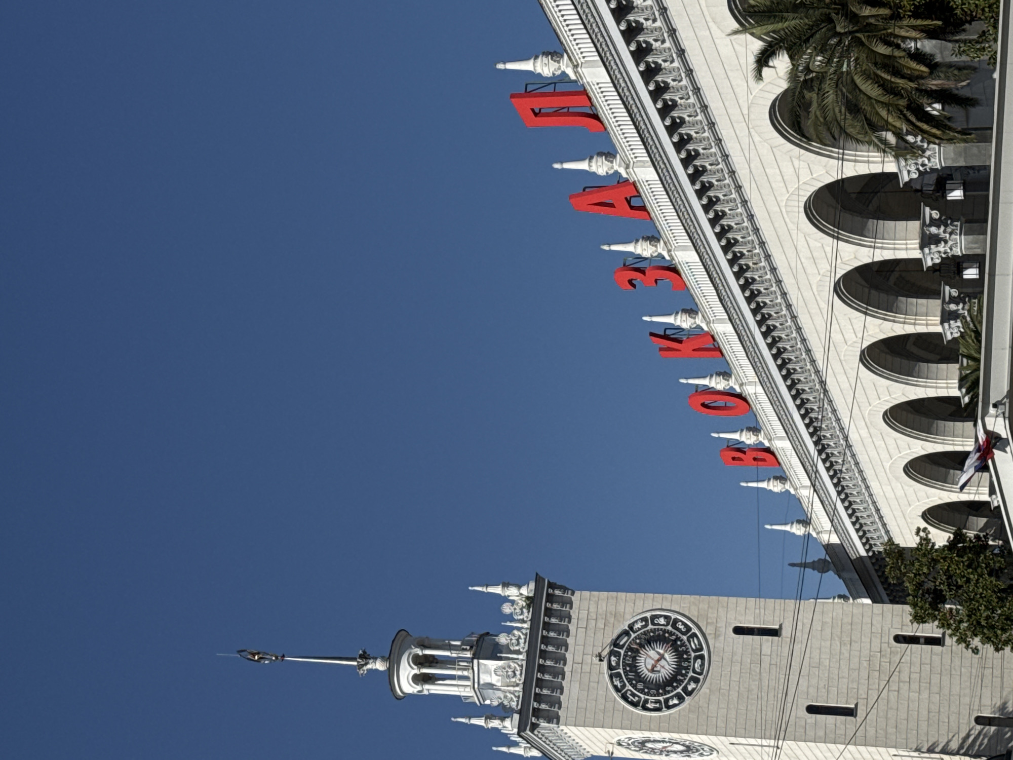 The image shows the Sochi Sea Port building in Sochi, Russia, under a clear blue sky. The architecture features a clock tower and a white façade with arched windows and red Cyrillic lettering on the top. A palm tree is visible adjacent to the building.
