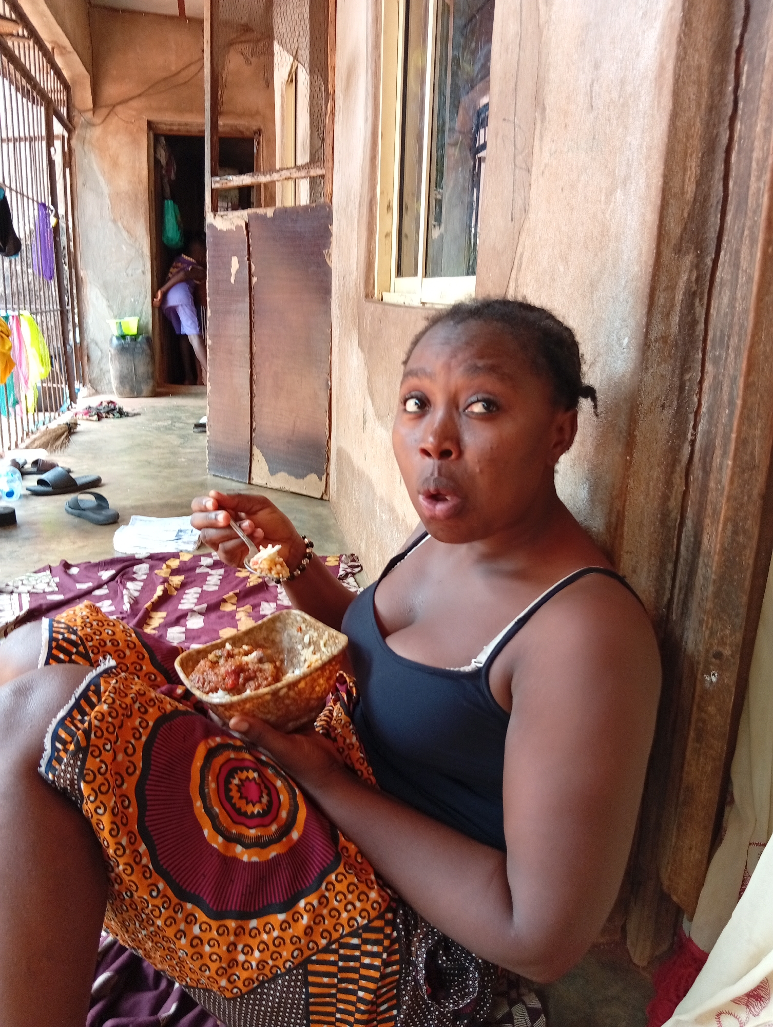 A dark-skinned woman sits, eating, with a surprised expression. She is in Ogidi, Nigeria, wearing a black tank top and a patterned orange and brown skirt, with a bowl of food in her lap and a spoon in her hand. Behind her is a concrete wall with a window, and to the left is an open doorway leading to an interior space with miscellaneous items. Clothes are draped on a railing.