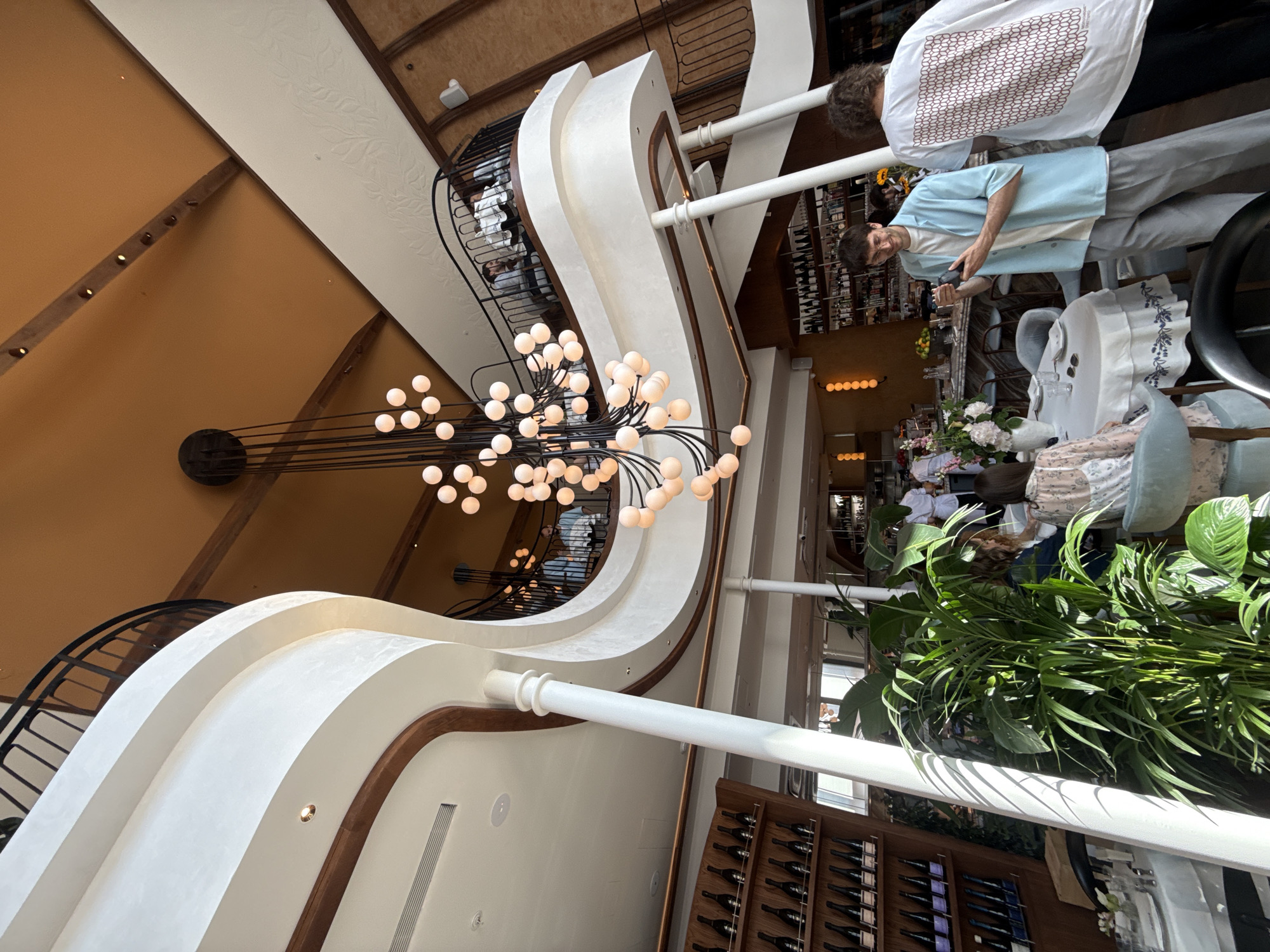 The image depicts the interior of a multi-level restaurant with a grand chandelier and ornate balconies. The lighting suggests it is daytime, with natural light streaming in. In the foreground, a large potted plant sits near tables set for dining. Two individuals are prominent: one man in a light blue shirt and grey pants is holding a phone, and another person in a white t-shirt with a patterned design stands with their back to the camera. The restaurant features rows of wine bottles on shelves and a bar area in the background. The architectural style appears modern with curved lines and elevated walkways. The city of Moscow is not visible in this interior shot.
