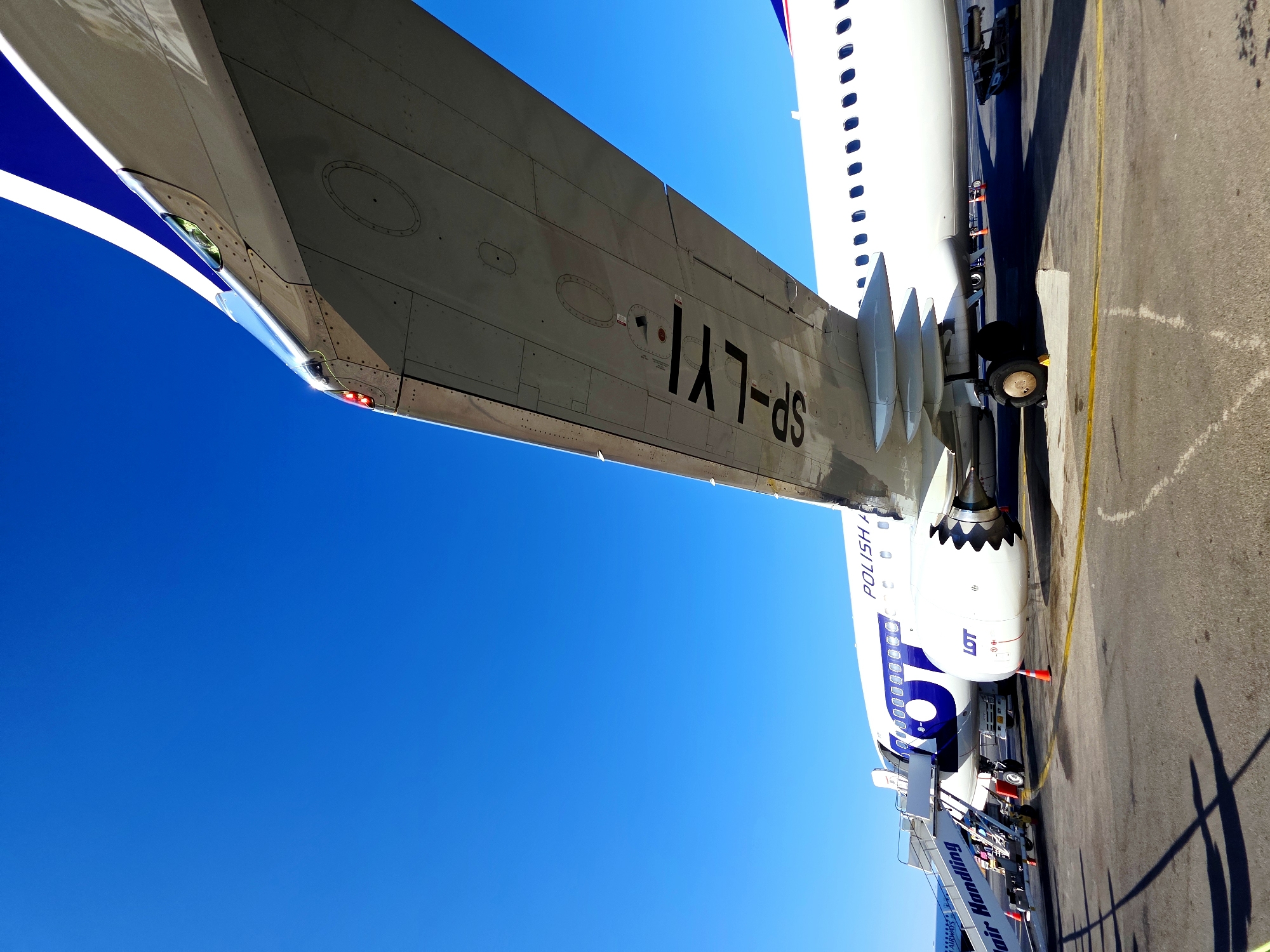 The image depicts a white LOT Polish Airlines aircraft parked on an airport tarmac under a clear blue sky. The perspective is from ground level, looking up towards the underside of the wing and the fuselage. The aircraft's registration number, SP-LXi, is clearly visible on the underside of the wing. A set of stairs is positioned adjacent to the aircraft's door, and the text "Air Handling" is visible on the stairs. In the background, another aircraft is partially visible. The tarmac is marked with yellow lines, and traffic cones are placed around the aircraft. The scene appears to be set at an airport in Nea Alikarnassos Municipal Unit, Greece, suggested by the presence of the LOT Polish Airlines aircraft which frequently flies to Greek destinations. There are no people visible in the image.
