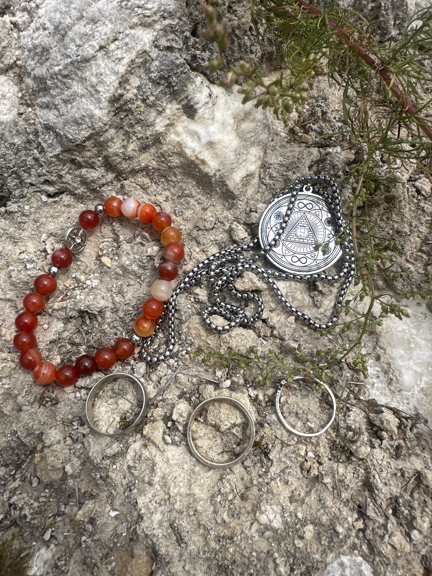 A close-up, high-angle shot shows a collection of jewelry items laid out on a textured, rocky surface. The jewelry includes a bracelet made of carnelian beads with a silver charm, a silver chain necklace with a round pendant featuring a triangular design and infinity symbols, and three silver rings. Green foliage is visible in the upper right portion of the frame. The scene appears to be outdoors, with the natural texture of the rock and plant life suggesting an outdoor environment. There are no people or visible actions occurring. The image is devoid of any context or clues that would place it specifically within the city of Simskii, Russia.