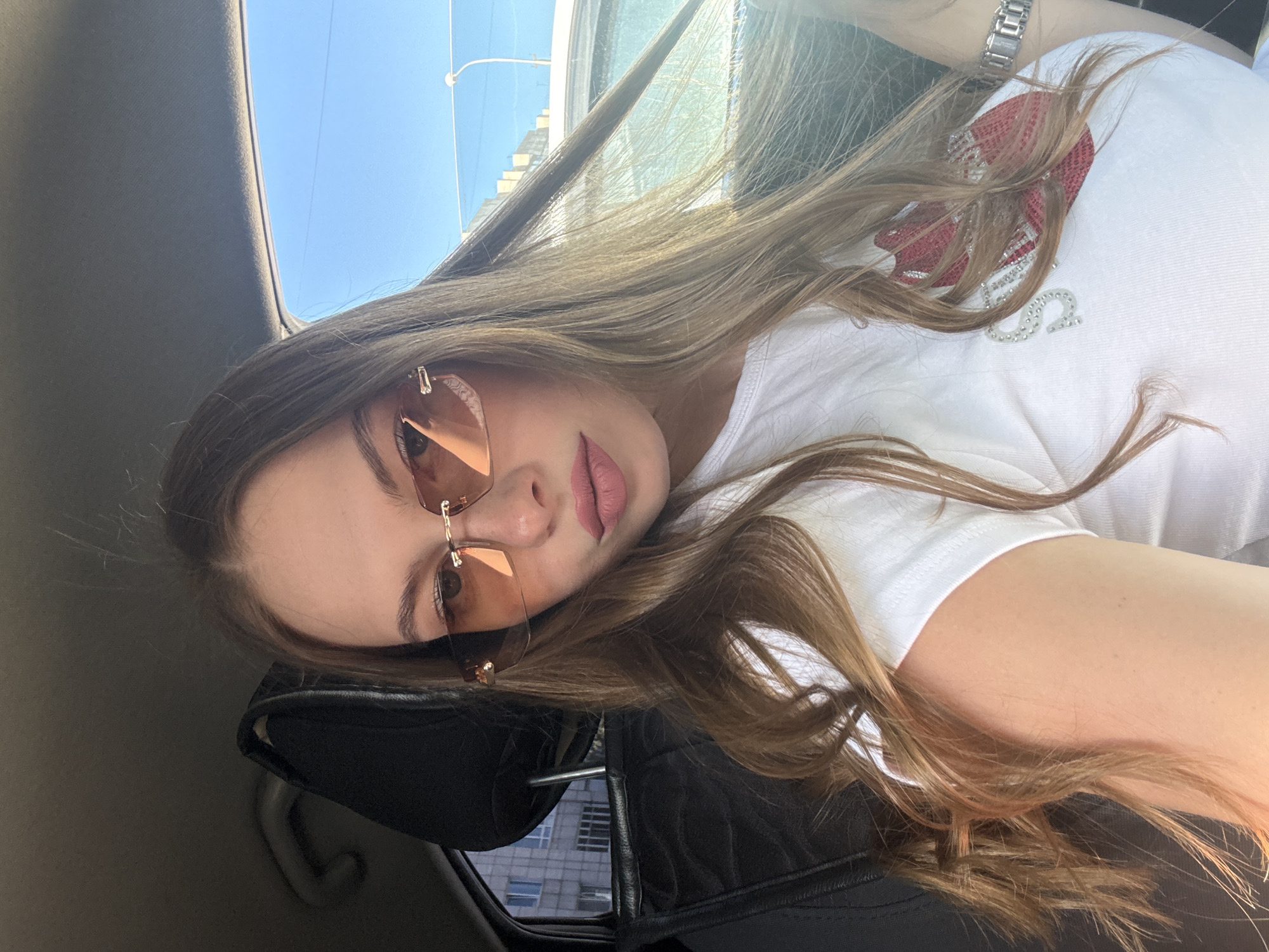 A young woman with long, light brown hair and light brown eyes is sitting in the passenger seat of a car. She is wearing rimless, rose-tinted sunglasses and a white t-shirt with a red lip design adorned with rhinestones. Her makeup includes pink lipstick. The car interior is visible, with black leather seats and a headrest. Outside the car window, buildings and a lamppost suggest an urban environment. The sky is clear and blue. This image was taken in the city of Perm, Russia.