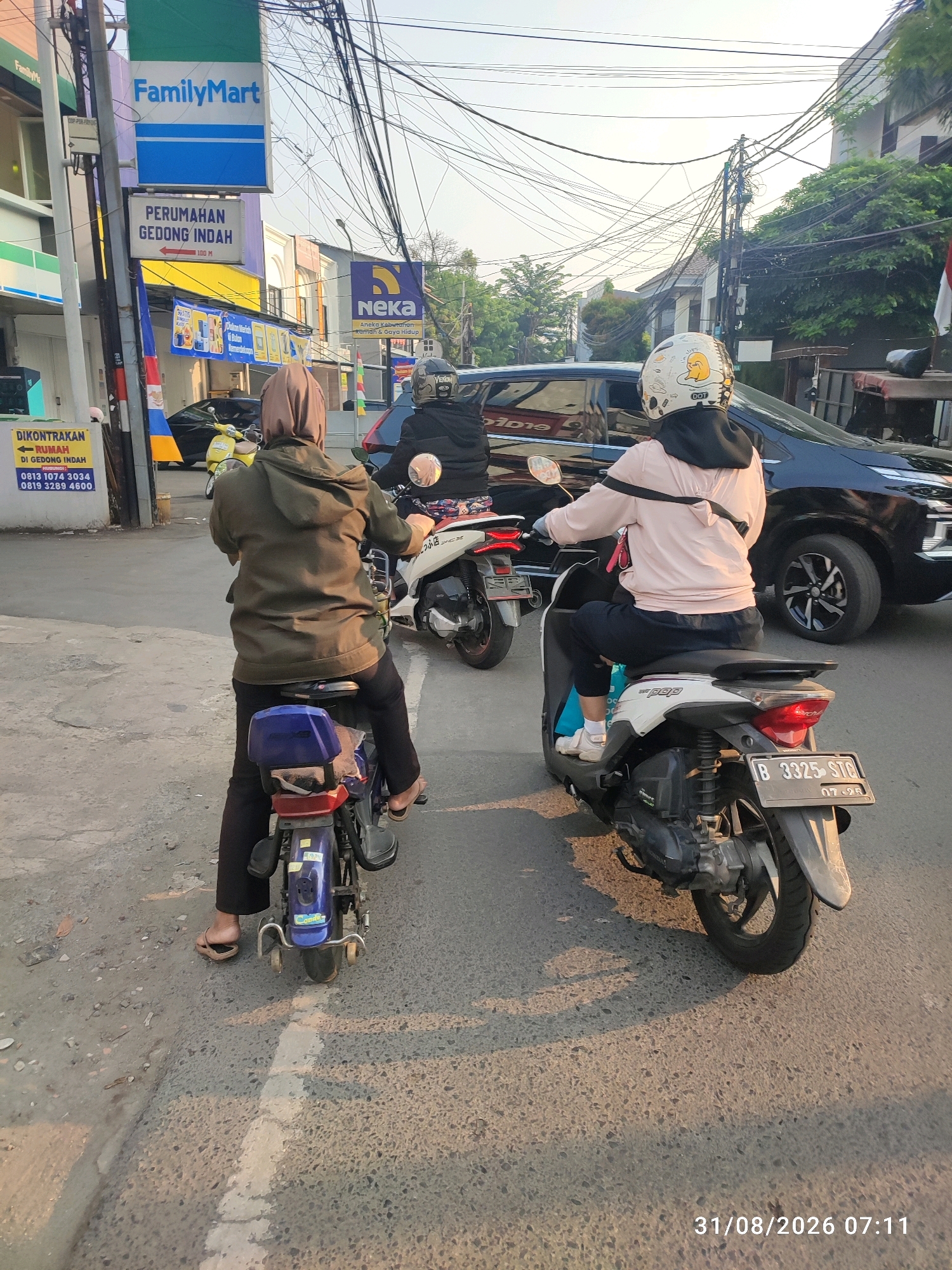 The image captures a street scene in East Jakarta, Indonesia, likely in the vicinity of Gedong Indah. Two individuals on motorcycles are visible, with the foreground dominated by a person wearing a brown hooded jacket and a hijab, riding a blue motorcycle. They are looking forward, and their legs are clad in black pants and brown sandals. Behind them, another person wearing a light pink jacket and a black hijab is riding a white motorcycle. This individual has a helmet decorated with cartoon characters, including what appears to be Winnie the Pooh. Their legs are in black pants and white sneakers.
In the background, several vehicles are present, including a black SUV to the right of the image and a yellow motorcycle further back. A large number of electrical wires crisscross the sky, indicating a densely populated urban area. Signs are visible on the left side of the image, one of which reads "FamilyMart," a convenience store chain. Another sign, in Indonesian, indicates "PERUMAHAN GEDONG INDAH," suggesting the name of a residential area, and shows an arrow pointing left with "100 M." A sign for "NEKA" is also present. The overall environment appears to be a street with buildings lining one side and trees and houses on the other. The lighting suggests it is daytime, with shadows cast on the road. The image is timestamped "31/08/2026 07:11."