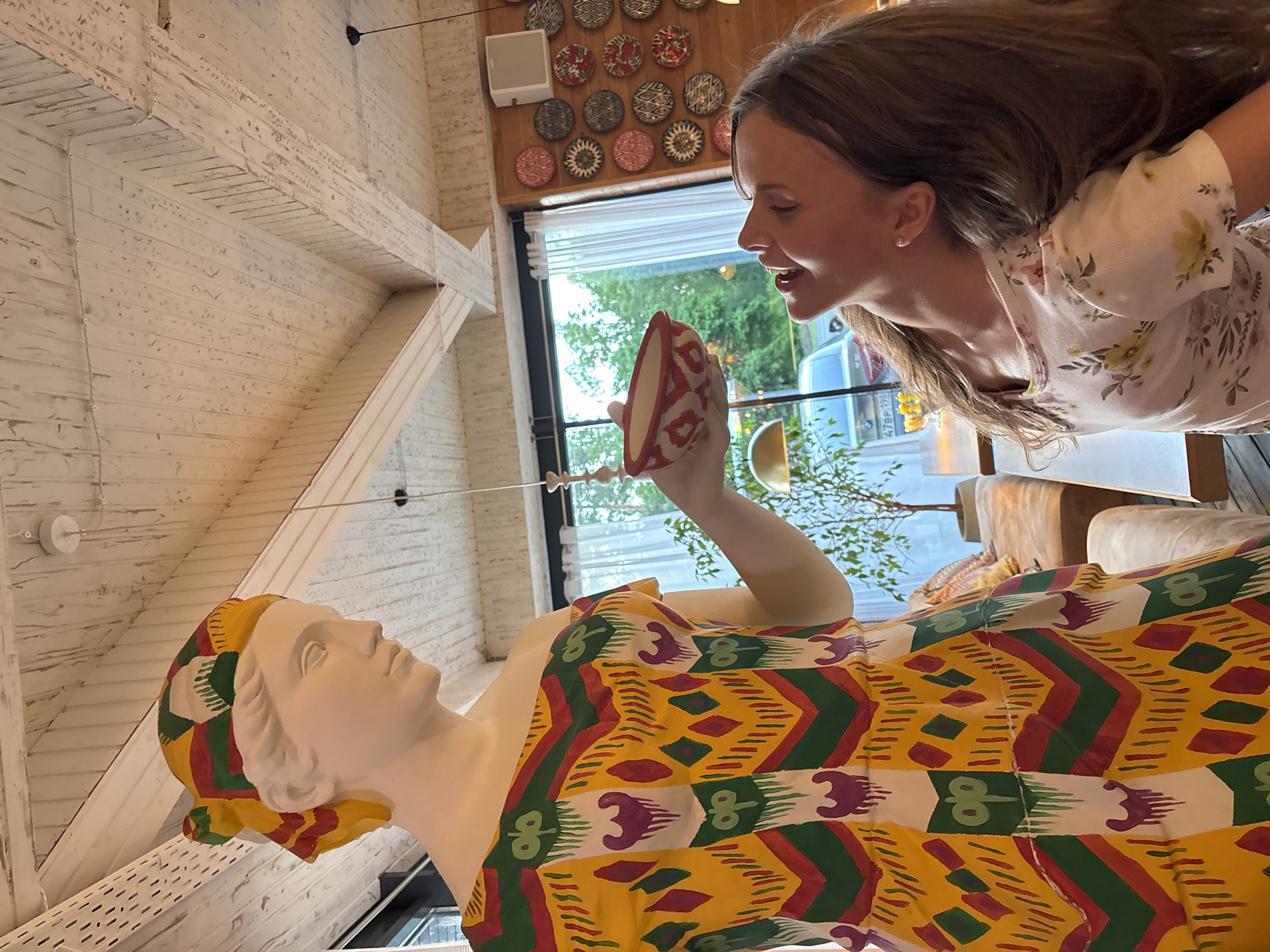 In Solnechnoye, Russia, within a brightly lit establishment featuring a high, angled ceiling with exposed wooden beams and white planking, a life-sized white statue of a woman stands prominently. The statue is adorned with a colorful headscarf and a vibrant yellow dress featuring a geometric pattern of red, green, and white zigzags and diamond shapes, interspersed with stylized figures. In its outstretched hand, the statue holds a small, red and white patterned bowl.
Interacting with the statue is a young Caucasian girl with long, brown hair, wearing a light-colored t-shirt with a floral print. Her head is tilted back, her mouth open in a laugh or exclamation, and her eyes are directed upwards towards the statue. The scene appears to be set in a cafe or restaurant, with a large window in the background revealing trees and a parked car. The wall behind the girl is decorated with a collection of round, patterned plates. In the mid-ground, a plush sofa and a potted plant are visible, suggesting a comfortable seating area. A metallic lampshade hangs from the ceiling. The overall ambiance is lively and playful, with the girl seemingly engaged in a whimsical interaction with the statue.