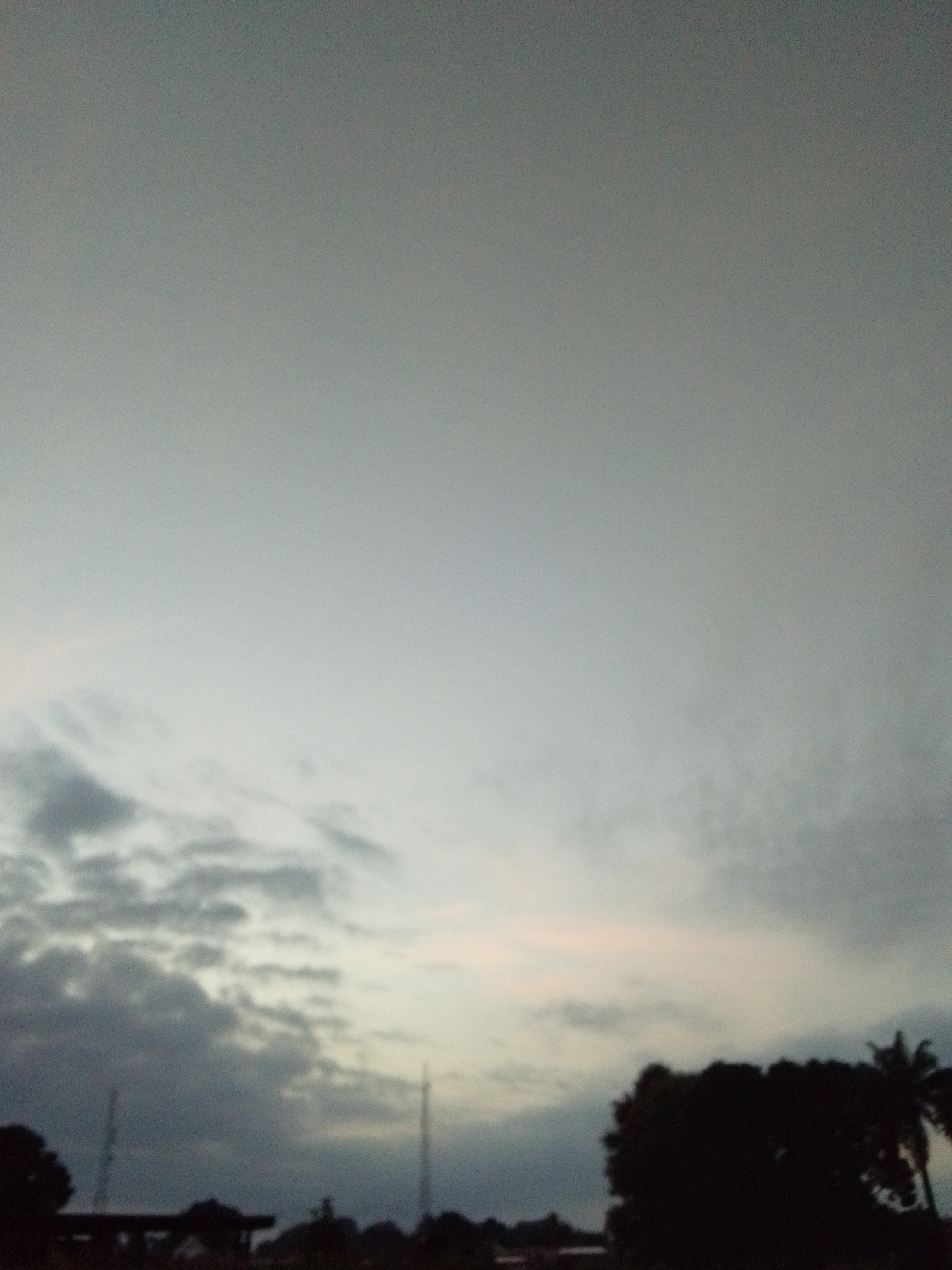 The image depicts a hazy, overcast sky over a silhouetted landscape. The sky occupies the majority of the frame, transitioning from a lighter hue near the horizon to a darker, grayish tone at the top. Wispy clouds are visible, particularly in the lower half of the sky, with some appearing darker and more defined than others.
The lower portion of the image reveals a dark, indistinct silhouette of trees and structures. On the far right, the silhouette of a palm tree is visible. Two thin, vertical poles, possibly antenna towers, are present in the middle ground. The overall impression is that of an outdoor scene at dusk or during a cloudy day. No people or specific actions are discernible. The location is identified as Serti, Nigeria.