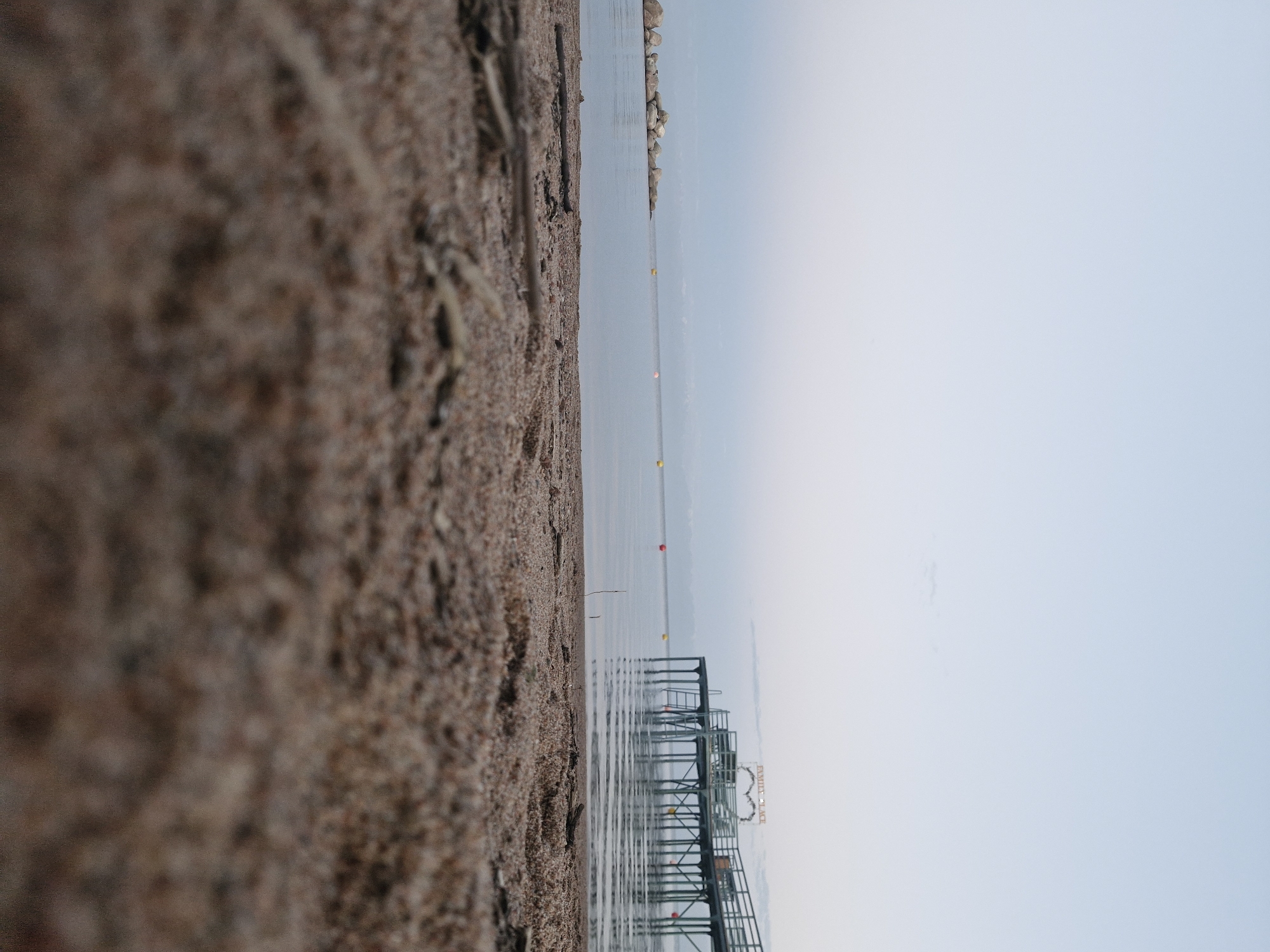 This is a low-angle, wide shot of a beach and a body of water at dusk or dawn. The foreground is dominated by dry sand with some small twigs and debris scattered across it. In the mid-ground, the calm water of a lake or sea is visible, with a row of large rocks on the left side and several yellow and red buoys in the distance. On the right side of the mid-ground, a wooden pier extends over the water. The pier has railings, support posts, and a sign at the end that reads "FAMILY PLACE". A heart-shaped decoration is also visible on the pier. The sky is clear and a pale blue, with a gradient fading to white near the horizon. The location is identified as Bulan-Segettu, Kyrgyzstan. There are no people or animals present in the image.