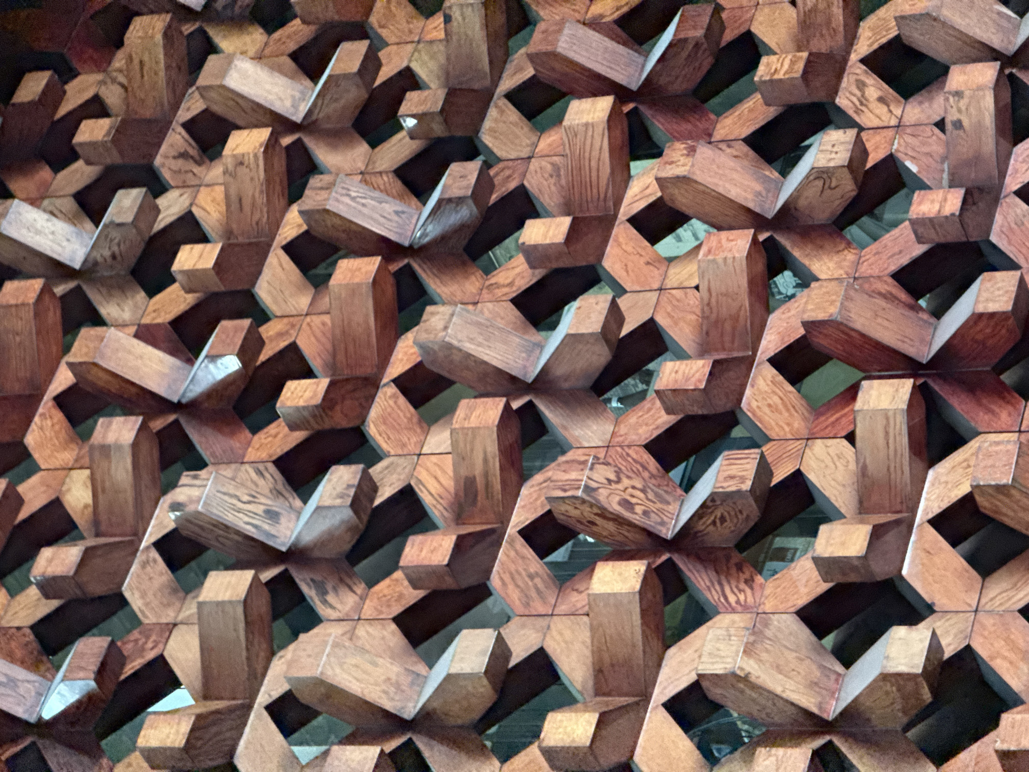 The image displays a close-up view of an intricate, three-dimensional wooden screen or wall panel, likely an architectural or decorative feature in Mexico City, Mexico. The primary subject is the complex geometric pattern formed by numerous interlocking wooden blocks. These blocks are crafted from wood with a warm reddish-brown hue, showcasing distinct grain patterns and variations in color, from lighter tan to deeper reddish-brown.
The design features a repetitive arrangement of hexagonal and star-like shapes, creating a highly textured surface. Individual wooden pieces are precisely cut and fitted, some appearing to recede into the background, while others protrude forward, casting shadows and enhancing the sense of depth. This volumetric arrangement creates a dynamic interplay of light and shadow across the entire surface. The gaps within the pattern reveal a darker background, which further emphasizes the three-dimensionality of the wooden elements.
No activity or event is taking place; the image captures a static architectural detail. The consistent lighting suggests either artificial illumination or diffused natural light, highlighting the varying planes and angles of the wooden structure. No people or visible text are present in the image. The intricate craftsmanship and geometric complexity suggest a detailed and artful decorative installation.