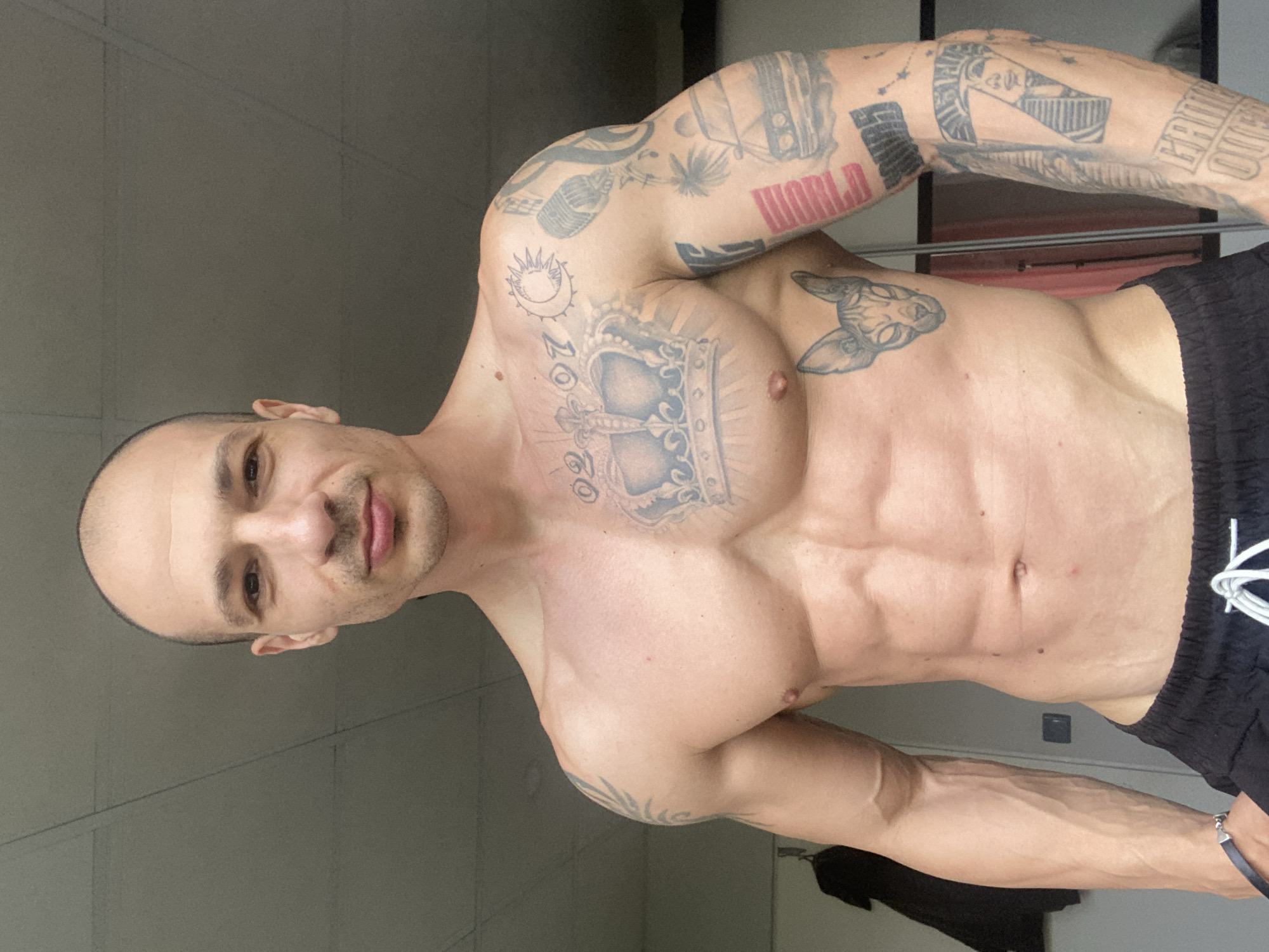 A muscular, light-skinned man with a shaved head poses shirtless, showcasing his torso, shoulders, and arms which are covered in tattoos. He wears dark shorts with white drawstrings. The background appears to be an indoor setting with a pale green wall and a dark object possibly clothing hanging in the lower area of the shot. There is no indication of Pleven, Bulgaria.