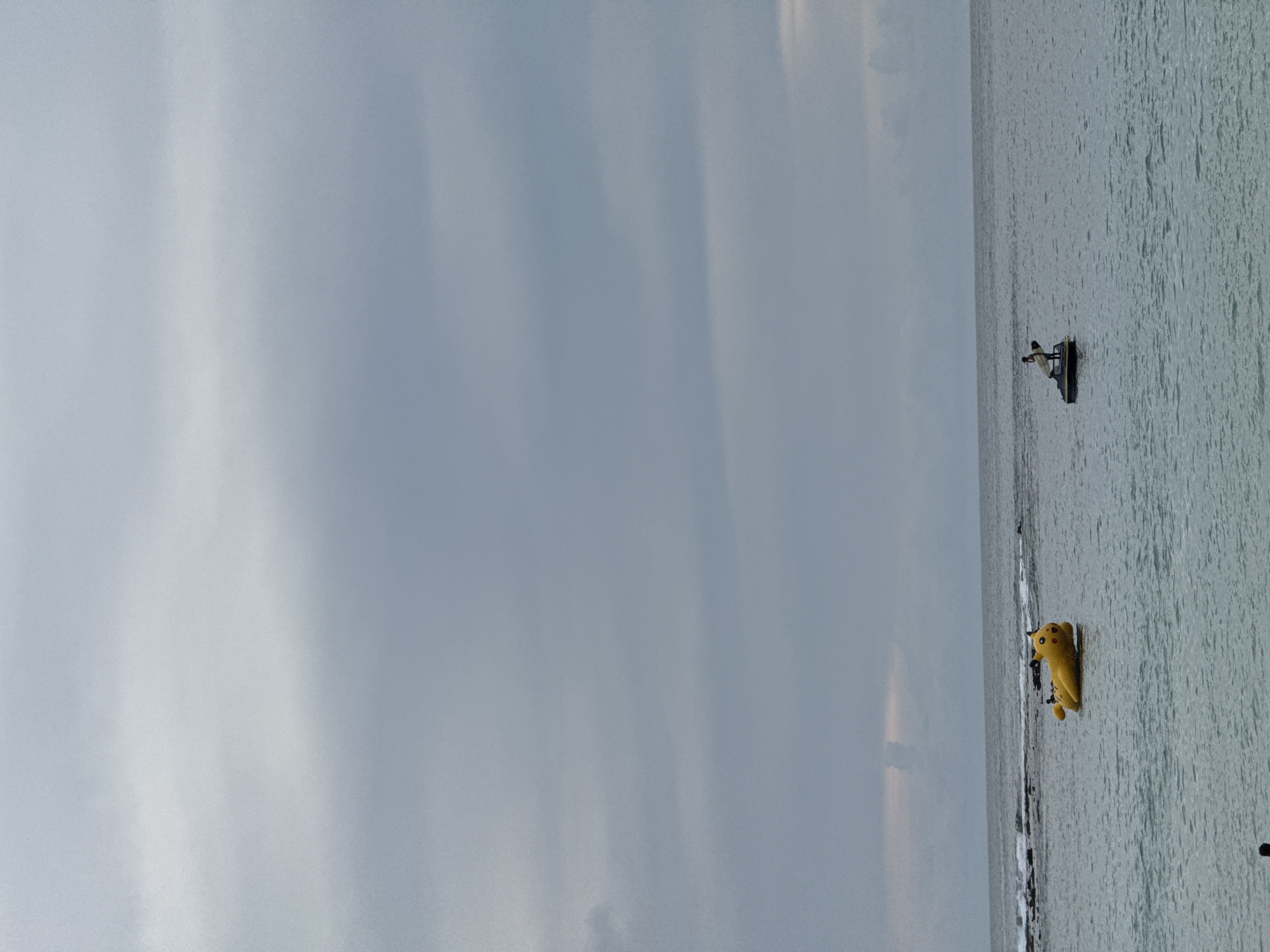 An overcast sky hangs over a body of water where a yellow inflatable Pikachu floats on the left side, and a jet ski with a person holding a surfboard is on the right side. The Pikachu appears to be a large inflatable toy, possibly for recreational use on the water. The jet ski is positioned further out in the water, with a solitary figure standing on it, holding a surfboard. The environment suggests a coastal or marine setting, likely a beach or open sea area, given the presence of waves and the vast expanse of water. The overall mood is calm and somewhat subdued, consistent with the cloudy weather. There are no clear indications of the specific city of Meunasahbale, Indonesia within the image itself, as it is a scene of water and sky with recreational watercraft.