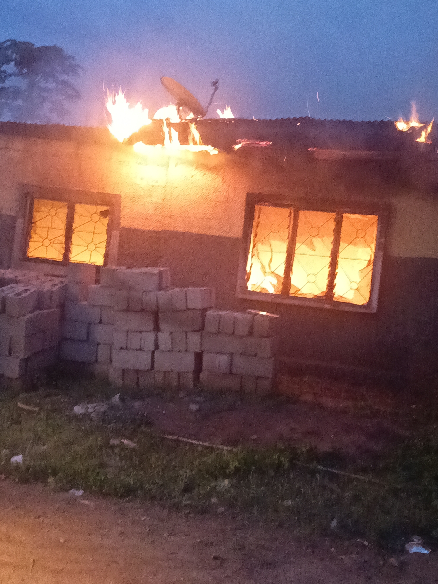 The image captures a house on fire in the city of Akwanga, Nigeria. Flames are visible engulfing parts of the roof, and smoke is rising into the twilight sky. Two windows are illuminated by the fire, suggesting the interior is also burning. In front of the house, stacks of concrete blocks are piled on the ground, possibly materials for construction or repair. The surrounding environment appears to be a dimly lit, possibly rural or semi-urban area, with grass and dirt visible in the foreground. The overall scene depicts an active fire incident.