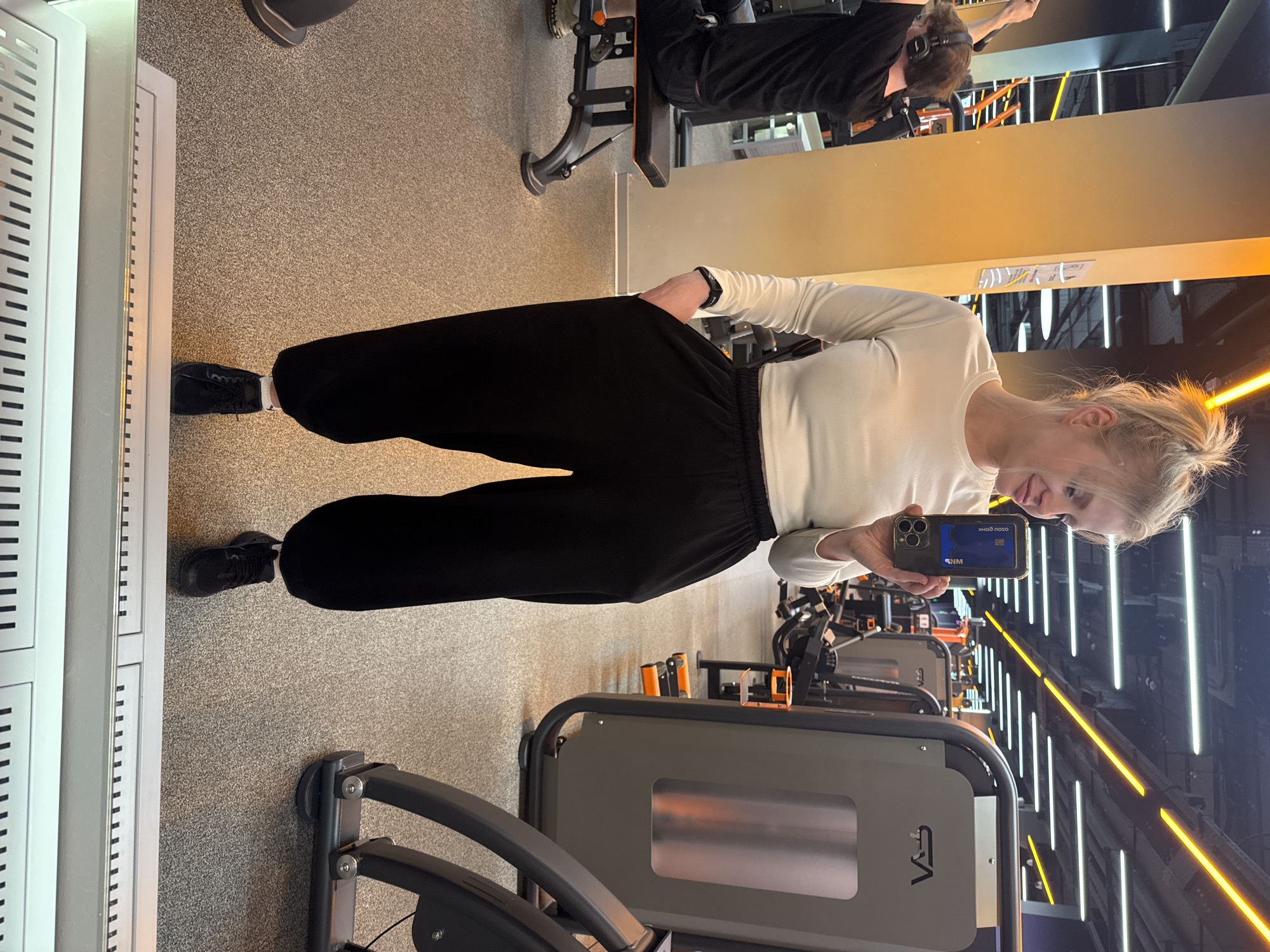 A gym interior in Saint Petersburg, Russia, features a blonde woman dressed in a white, long-sleeved shirt and black pants, taking a mirror selfie with a smartphone. Exercise machines fill the background, illuminated by bright, linear lighting. Another individual, possibly male, wearing dark clothing and headphones, is using a gym machine in the distance.
