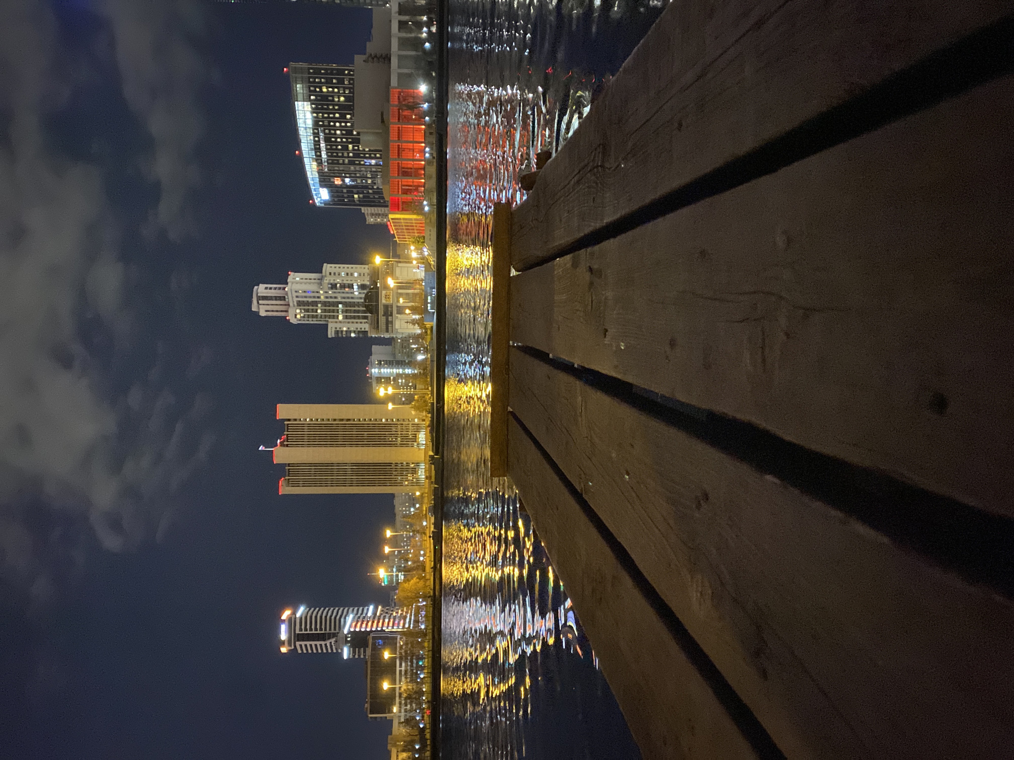 At night, a dock or walkway made of dark wood stretches into the water. The water reflects the lights of the city of Yekaterinburg, Russia. Several tall buildings are visible in the background, along with streetlights. The sky above is dark, with some clouds.