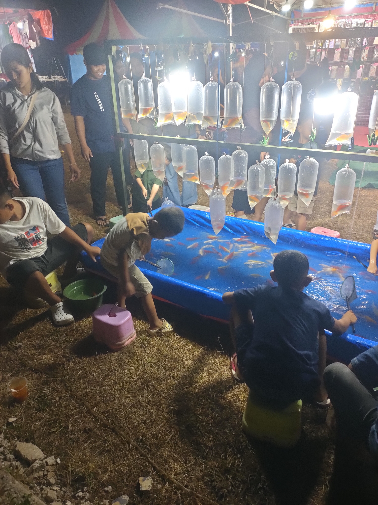 The image depicts a nighttime scene at an outdoor fair or market, likely in Subang, Indonesia, given the context. In the foreground, two young boys are positioned near a large, bright blue inflatable pool filled with numerous small, orange and white fish. One boy, wearing a dark t-shirt, sits on a yellow stool and holds a black fishing net, appearing to be actively trying to catch fish. The other boy, in a light brown shirt and shorts, is leaning over the pool. Around them, on the dry, grassy ground, are scattered debris and several plastic containers, including a green bucket and a pink stool.
Behind the pool, a row of clear plastic bags, each containing a few orange fish, are suspended from a metal rack under bright lights. Several adults and teenagers are visible in the background, observing the activity or browsing stalls that are partially obscured. One woman in a grey jacket and blue jeans stands to the left, while a young man in a dark t-shirt with white lettering is to her right, looking towards the fish. The overall atmosphere suggests a lively event, possibly a carnival game or a market stall selling fish. The lighting is artificial and somewhat harsh, creating strong shadows.