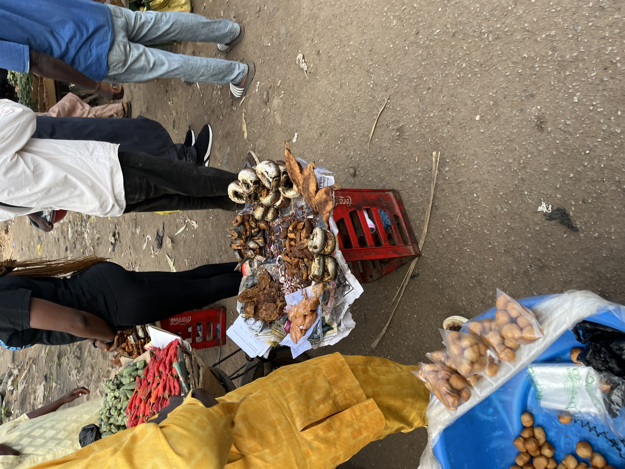 In Bukuru, Nigeria, a street vendor is selling fried fish and other food items from a stall. A woman in a black t-shirt and black pants stands behind the stall, facing to the right. In front of her, a variety of fried fish, including whole fish and fish pieces, are displayed on a newspaper-covered surface. Some small, round, fried items are also visible in a blue plastic basin, some of which are packaged in clear plastic bags. Red chili peppers and okra are also for sale on the stall. A person wearing a yellow traditional outfit is visible on the left side of the frame. Several other individuals, presumably customers and other vendors, are also present in the background, with some standing and others partially obscured. The ground is a dirt and gravel surface.