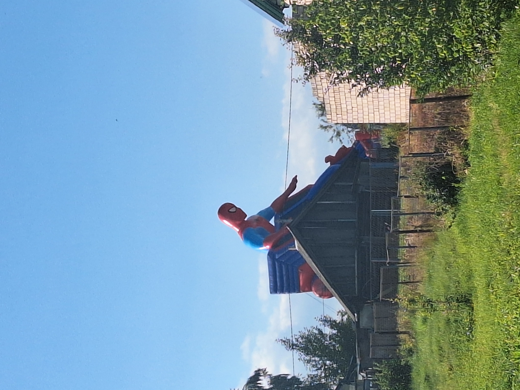 The image shows an inflatable Spider-Man figure perched on top of a wooden structure that resembles a small building with a pitched roof. The Spider-Man figure is red and blue, with its signature mask. The structure appears to be located in a rural or semi-rural area, with a fenced yard in the foreground filled with green grass and some vegetation. In the background, there are trees, a brick wall structure, and a clear blue sky. The overall scene suggests an outdoor recreational area or a residential property with a playful decoration. The image was captured in Блеўчыцкі сельскі Савет, Belarus.