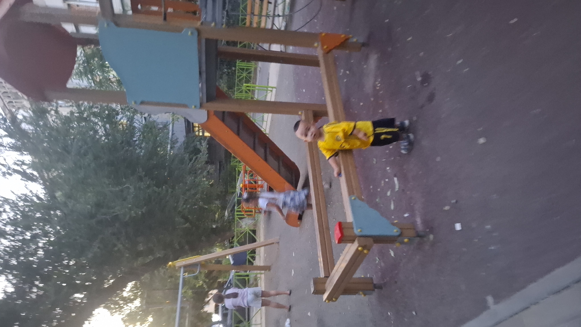 In Shymkent, Kazakhstan, a small child wearing a yellow t-shirt and black shorts with yellow stripes stands near a wooden playground structure. The child is looking towards the camera, with their right hand touching the wooden railing. In the background, a girl in a white shirt and patterned skirt is walking, and another woman in a white shirt and shorts is standing further back near a swing set. The playground features an orange slide, a red roof, and a blue panel on the structure. The ground is a dark reddish-brown, with scattered debris. Trees with green foliage and a building are visible behind the playground. The lighting suggests it is either early morning or late afternoon.