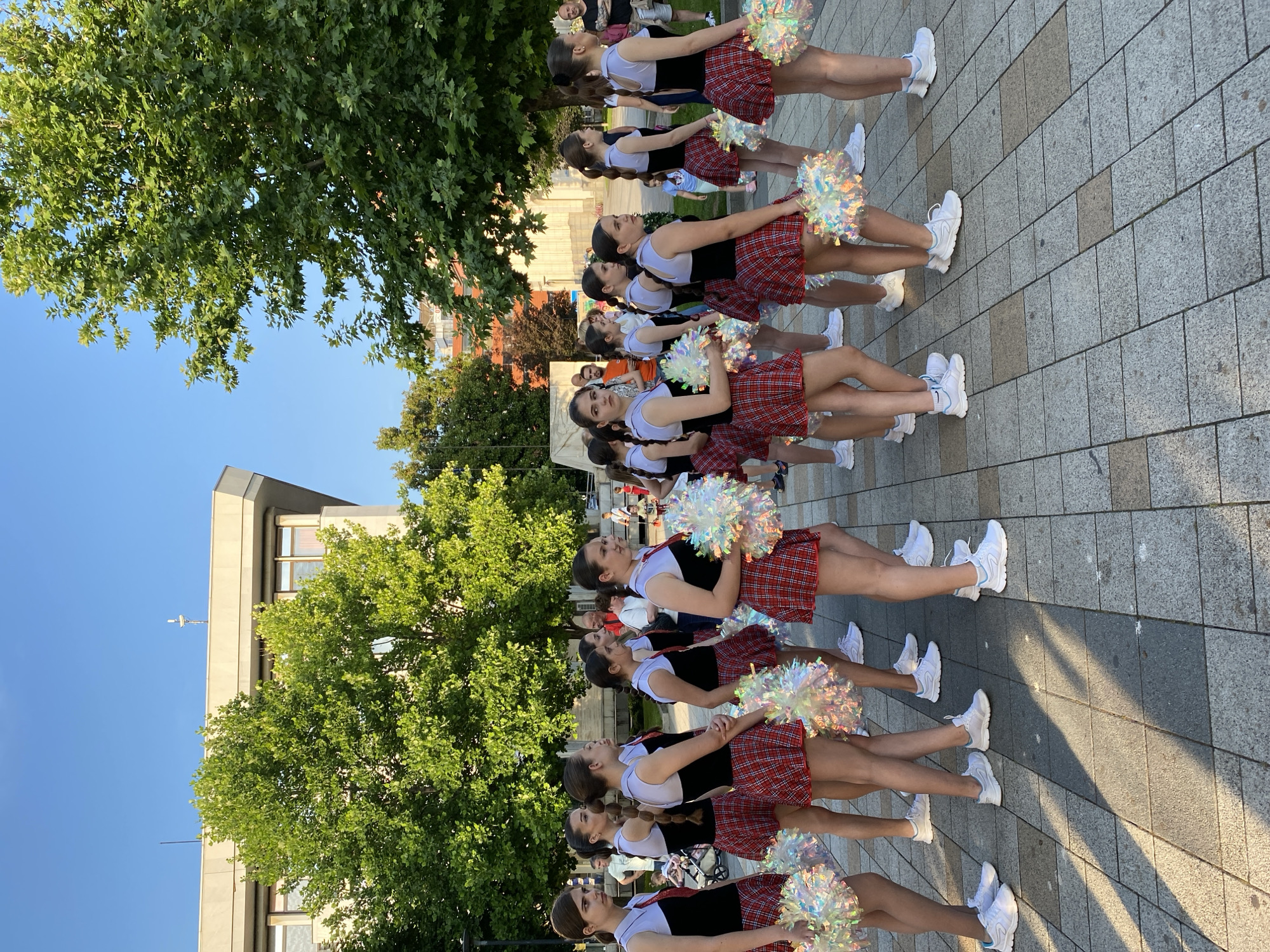 The image depicts an outdoor scene in the central pedestrian area of Pleven, Bulgaria, likely in front of the Regional Administration building, identifiable by its light-colored, multi-story facade and flat roof with antennas.
The foreground and mid-ground are dominated by a group of approximately 15 to 20 young females, appearing to be teenagers or pre-teens, uniformly dressed. Their attire consists of a sleeveless black top with a light grey or white upper panel, a white collar with a red necktie, and a red and black plaid (tartan) miniskirt. Each individual wears white athletic shoes, some with light blue accents, and holds one or two large, shimmering, iridescent pom-poms that appear predominantly white, silver, or light multi-hued. Most of the girls have dark hair styled in ponytails or braids. They are standing in multiple rows, generally facing towards the left side of the frame, with some looking forward or subtly towards the camera. Their expressions are mostly neutral or focused.
In the background, several adults and a few children are scattered. Adults are casually dressed, some observing the uniformed group, others appearing to walk through the area. A man in an orange t-shirt, another in a dark t-shirt, and a woman in white are visible. A baby stroller is present on the far left.
The environment is an urban public space paved with rectangular grey and darker grey concrete or stone pavers, showing distinct shadows from the sun, indicating bright daylight conditions. Numerous large, mature trees with dense green foliage are prominent in the background and mid-ground, suggesting a park-like or tree-lined setting within the city. A clear blue sky is visible above the trees and building. The overall scene suggests a public gathering or performance event.