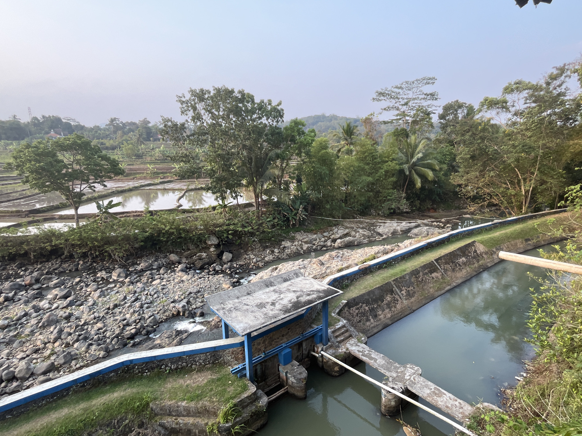 The image depicts a rural landscape in Krasak, Indonesia, characterized by agricultural fields and a water management system. In the foreground, a concrete structure with blue supports and a gray roof stands over a channel where water flows through. This structure appears to be a small dam or sluice gate, designed to control water levels. The surrounding area features a rocky riverbed with visible water and vegetation. Adjacent to this, a cultivated field, likely a rice paddy, is filled with water and reflects the sky. Further in the background, a dense arrangement of trees and foliage creates a lush green backdrop, with rolling hills visible in the distance under a hazy sky. The scene is natural and tranquil, showcasing a blend of agricultural land and water infrastructure.