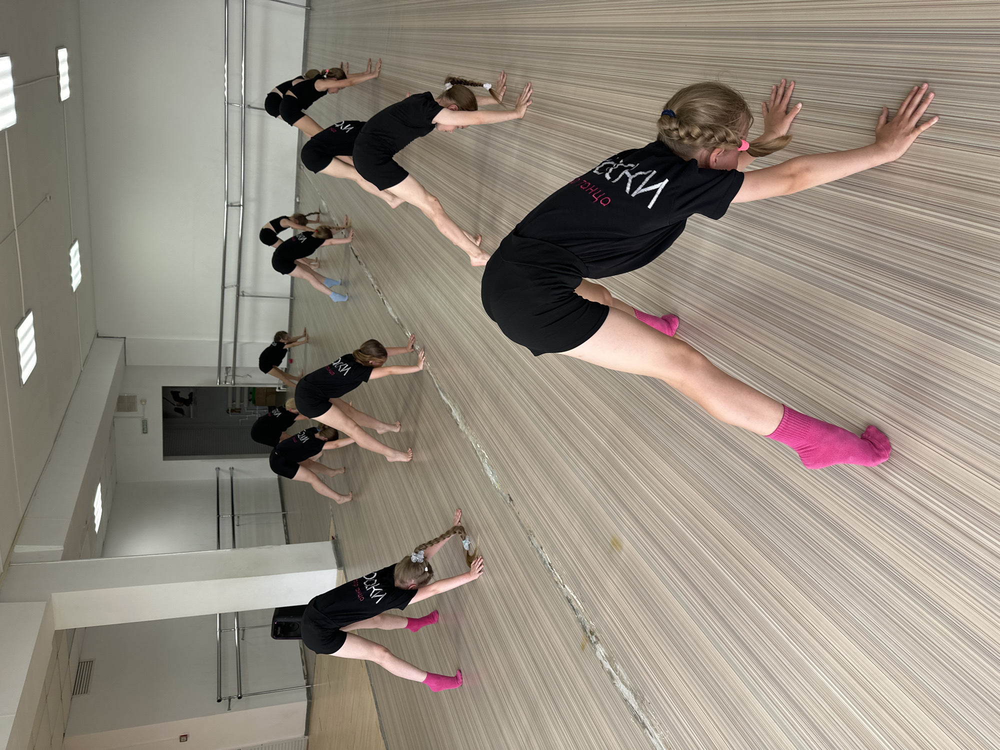 A dance class is in progress in Votkinsk, Russia. Ten young girls, all wearing black shorts and matching T-shirts with Russian text, are lined up, stretching against a wall that appears to be mirrored. Most of the girls are barefoot, but a few wear socks, including one pair that is bright pink. The studio features a wooden floor, ballet barres along one wall, and fluorescent lighting.