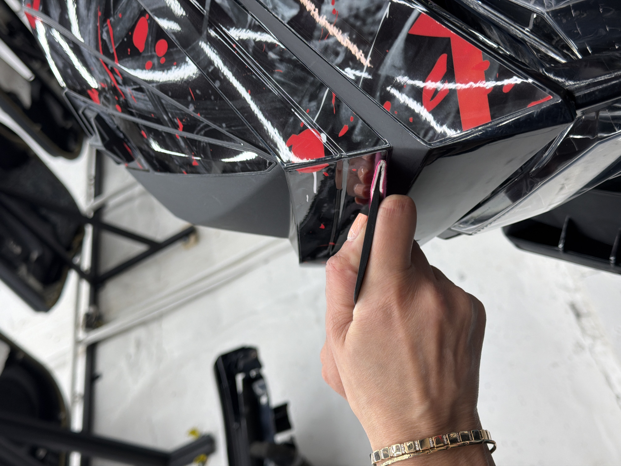 The image shows a close-up of a person's hand holding a black tool against a black and red patterned object, likely a car part. The object has a geometric design with glossy black surfaces and red splatters. The background reveals a workshop setting, with a white floor and metal frame structures supporting the car part. There is also a black object that appears to be a toolbox or machine in the background. The person is wearing a gold bracelet. There is no evidence in the image to confirm the location is in the city of Krasnoyarsk, Russia.