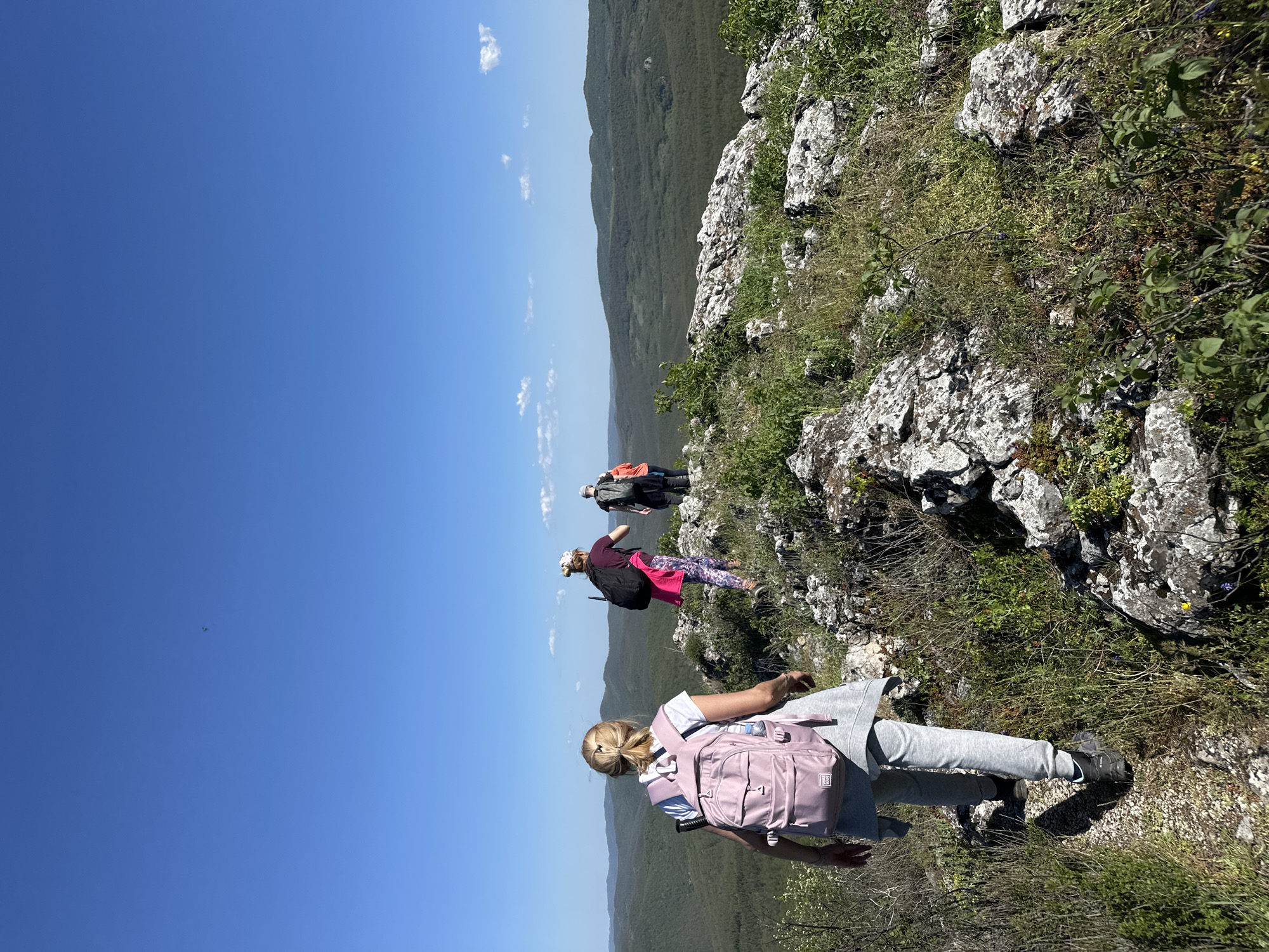The image shows a group of four individuals in a rocky, mountainous area, likely in the vicinity of Bilka, Bulgaria. The foreground depicts a person with a pink backpack, gray sweatpants, and dark-colored shoes, positioned on a rocky terrain covered with vegetation. Further in the distance, two more people are visible, one wearing a purple shirt and pink leggings, and another wearing an orange shirt. A fourth person is positioned in the distance. The landscape includes green hills and a clear blue sky with some scattered clouds. The setting appears to be a cliffside or a steep slope.