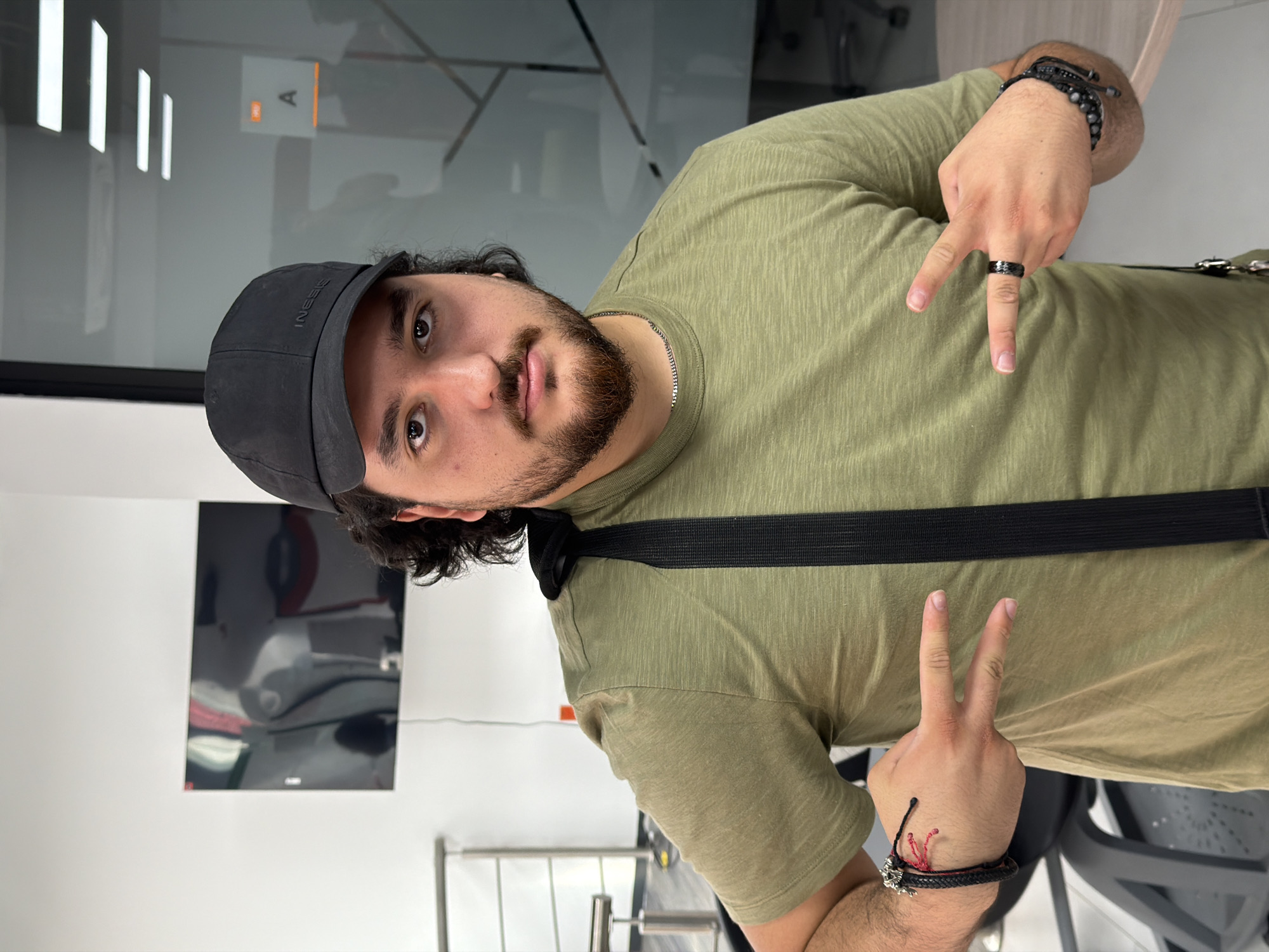 A man with dark hair and a beard is shown indoors. He wears a black cap, an olive-green T-shirt, and a black strap crosses his body. He makes hand gestures, holding up two fingers on one hand and pointing with one finger on the other. He wears a black ring and bracelets on both wrists.
A television is mounted on the wall behind him. There are lights on the ceiling and a doorway in the background. The scene appears to be in a gym or fitness-related establishment, given the visible gym equipment in the background.
While San Andrés Cholula, Mexico is mentioned, there is no clear visual evidence within the image to confirm this location.