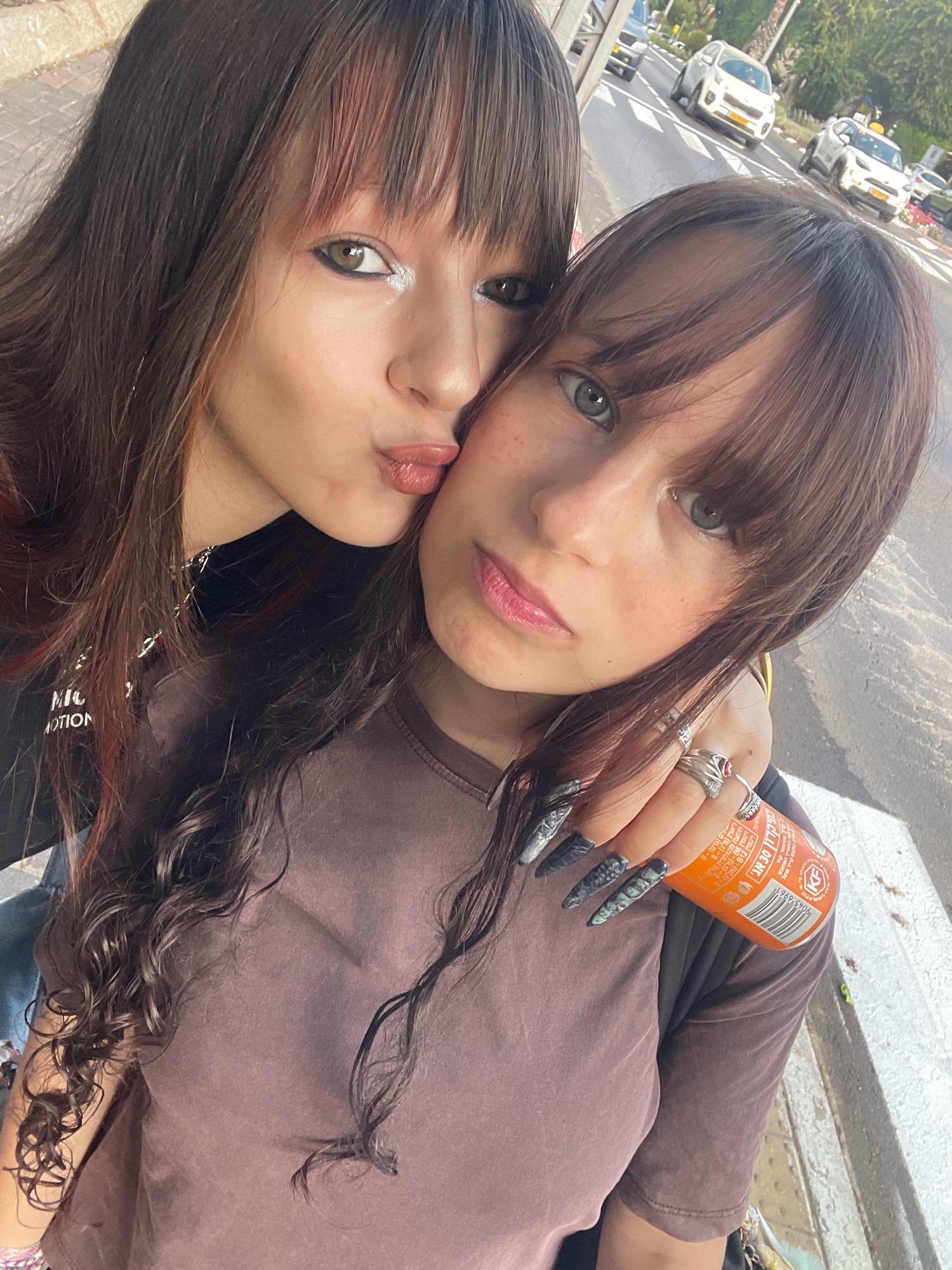 Two young women are posing for a selfie on a sidewalk in Raanana, Israel, with a street and cars visible in the background. The woman on the left is leaning towards the woman on the right, with her lips pursed as if to kiss her. She has long, dark brown hair with reddish undertones, bangs, and visible makeup, including winged eyeliner. She is wearing a black t-shirt with white lettering. The woman on the right has similar brown hair with bangs, light green eyes, and freckles on her cheeks. She is wearing a brown t-shirt and is holding an orange soda can in her right hand, which has long, dark, pointed nails with a textured design. The background shows a street with several cars, including white SUVs, and some greenery, consistent with a city environment in Raanana. The lighting suggests it is daytime.