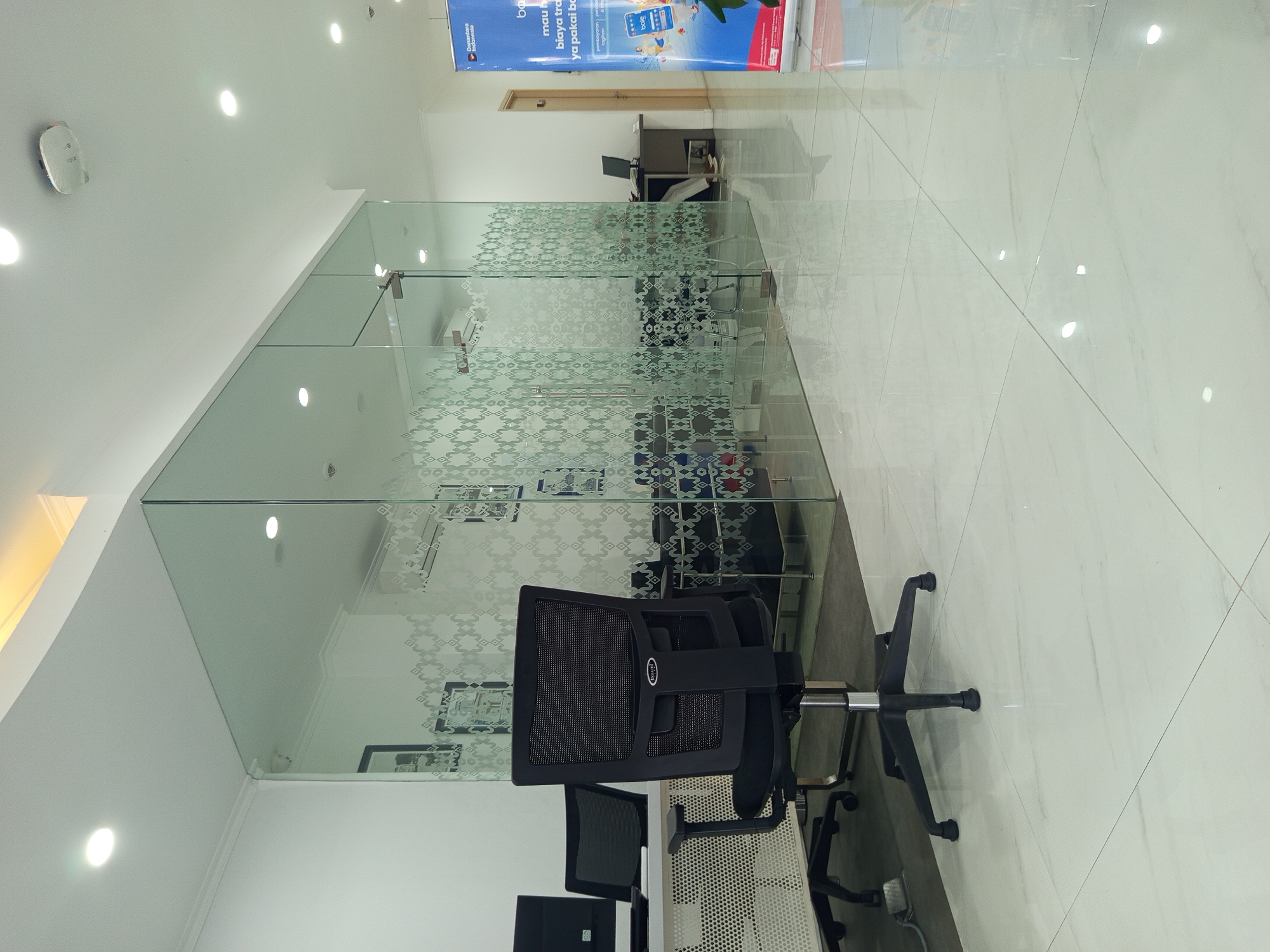 The image shows the interior of a modern office space, likely a bank or financial institution, given the banner visible on the right. The office features a large glass partition with a decorative pattern, separating different work areas. Inside the partitioned area, there are desks, chairs, computers, and what appears to be air conditioning units. In the foreground, a black office chair is prominently displayed on a polished, reflective tiled floor. The room is well-lit by ceiling lights. The banner on the right displays "D Nusantara Indonesia" and appears to be an advertisement for financial services. The overall impression is one of a clean, organized, and contemporary workspace. The location in Ardirejo, Indonesia is not discernible from the visual elements alone.