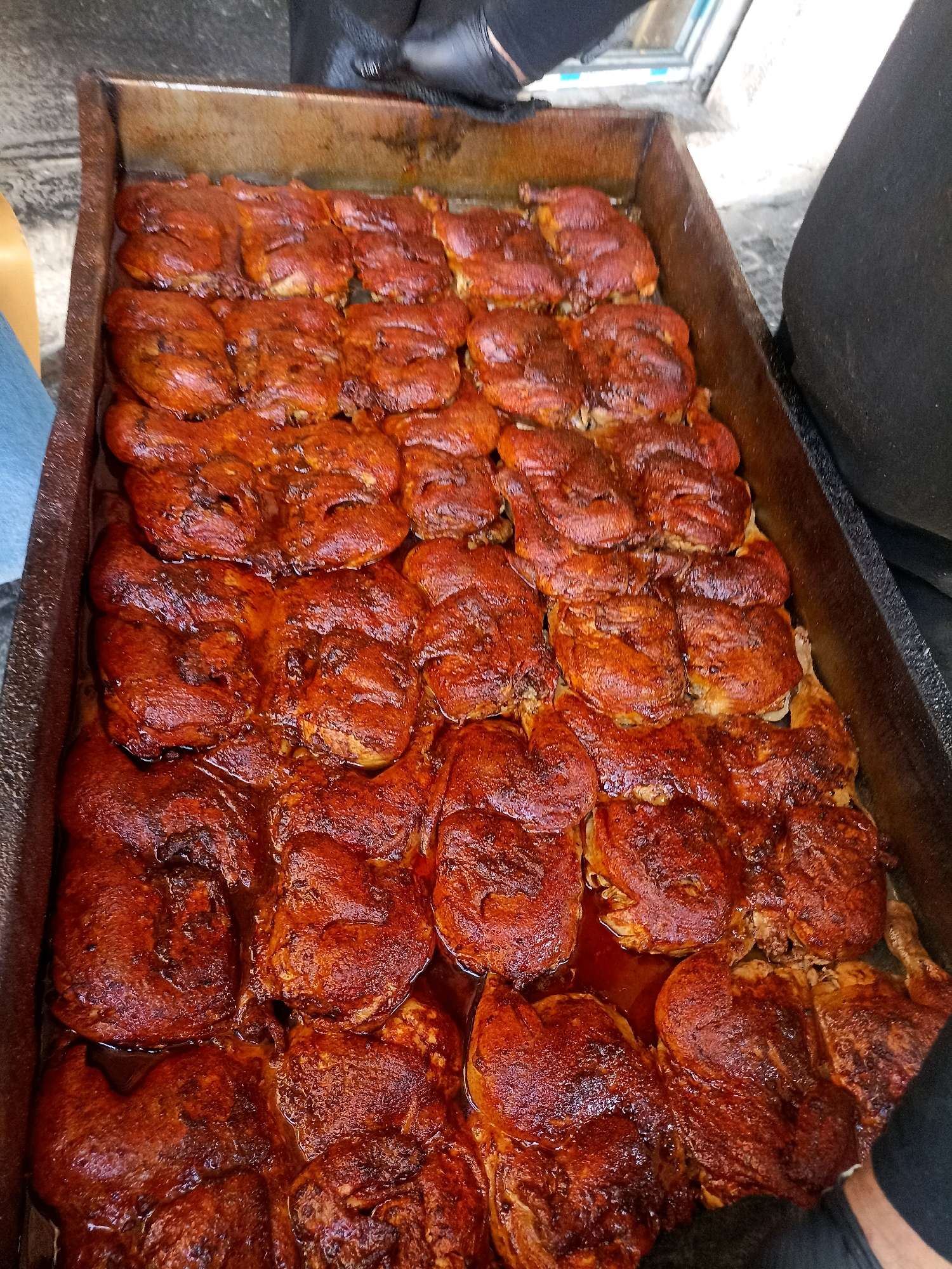 An overhead shot shows a large, rectangular metal pan filled with what appears to be cooked chicken pieces coated in a dark red spice rub. The chicken is arranged in rows, with some pieces overlapping. The pan itself is weathered and shows signs of use. In the background, a person's arm and shoulder, clad in black clothing and gloves, are visible on the right side, suggesting they might be handling the food. The scene appears to be outdoors, with a blurred backdrop that could be a street or market environment. The overall impression is of a food preparation or serving area, likely in Gaza, Palestinian Territories, given the context provided.