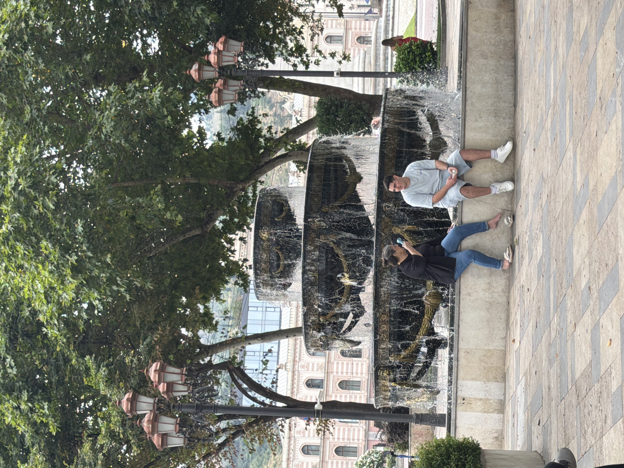 In Tbilisi, Georgia, two individuals are seated on the ledge of a multi-tiered fountain. A woman on the left, wearing a black jacket and blue jeans, holds a light blue object to her face, possibly a phone. A man on the right, in a light gray t-shirt and shorts, sits with his legs extended, holding a white object. The fountain, decorated with ornate gold accents, features water cascading down its dark tiers. Behind the fountain, large trees with lush green foliage dominate the background, with classic European-style buildings and a modern glass-fronted structure visible further beyond. Traditional street lamps with multiple globes stand to the left and right of the fountain. The ground is paved with a pattern of square tiles.