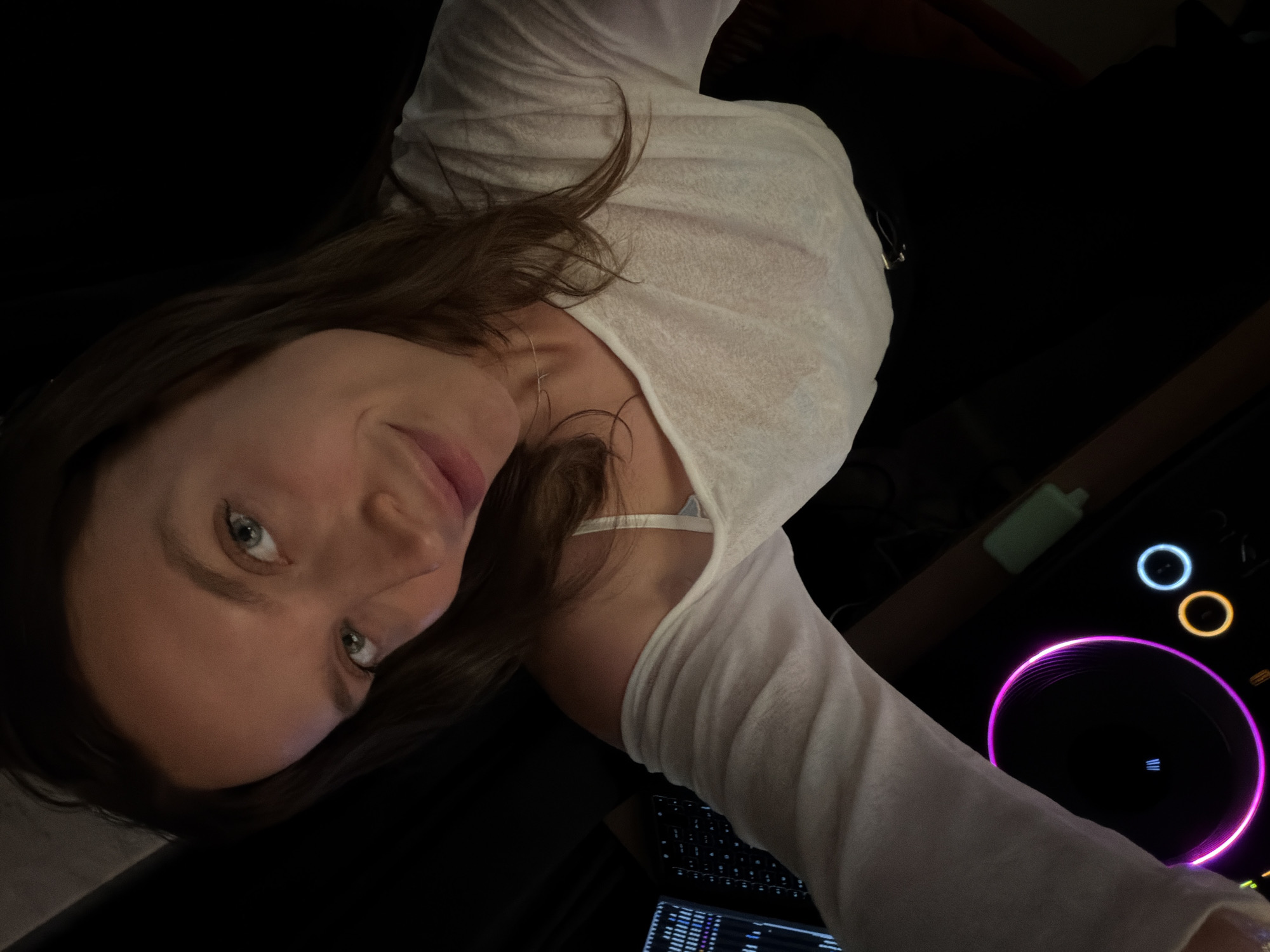 A high-angle selfie captured in a dimly lit indoor setting features a young adult female looking directly into the camera. She has long, dark brown hair and light green eyes, with a slight smile on her lips. She is wearing a loose-fitting, light-colored long-sleeved top, partially off her right shoulder, revealing a white bra strap. A delicate, thin gold-colored necklace with a small pendant is visible on her neck. Her lower body is obscured by dark clothing.
To the left of the subject, a vertical digital display with illuminated bars is partially visible. Below her right arm, electronic music equipment is prominent, featuring a large circular jog wheel glowing magenta and containing a small blue vertical light. Adjacent to this, two smaller circular lights glow orange and light blue. A small, light green, rectangular object rests nearby. The background is predominantly dark, suggesting an evening or nighttime environment, likely a studio or DJ booth. There is no discernible visual information within the image to confirm the location as Budva, Montenegro.
