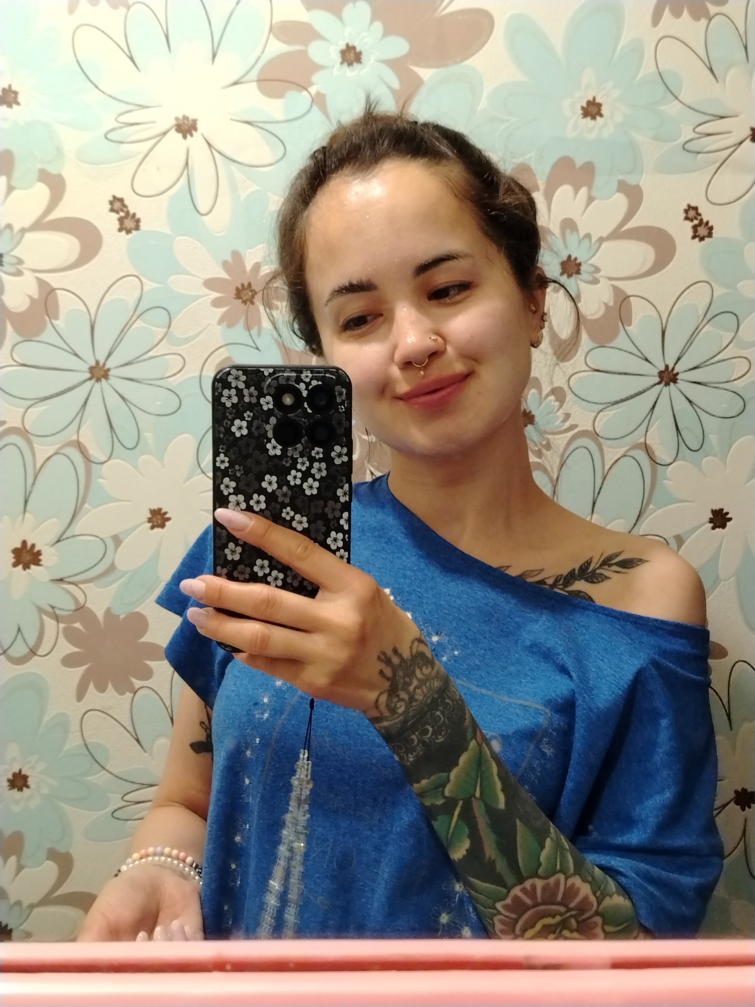A woman is taking a selfie in a bathroom. She has brown hair pulled back, light skin, and wears a blue off-the-shoulder shirt. Her left arm has a full tattoo sleeve, and she has a leafy tattoo on her right shoulder. She is holding a black phone with a floral case, displaying her long, manicured nails. She has multiple piercings in her ears and a septum piercing. The bathroom has floral wallpaper and a pink countertop. There is a beaded bracelet on her left wrist. The location is Гожан, Russia.