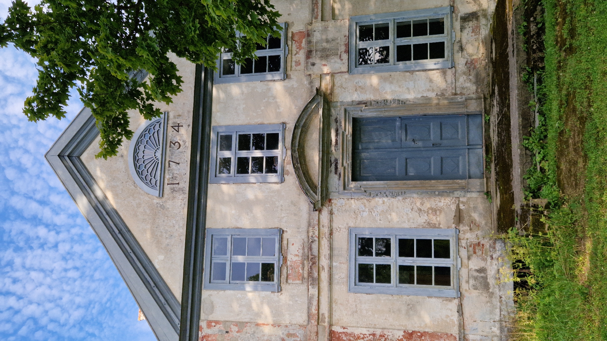 The image shows the exterior of an old, two-story building with peeling beige and red-toned plaster. The building has a triangular gable roof with grey trim. Above the central entrance, the year "1734" is inscribed below a decorative semicircular fanlight. There are several windows with light blue frames, each divided into six panes. A dark blue double door is centrally located on the ground floor, accessed by a set of stone steps. The steps and the ground around the entrance are covered in moss and overgrown with grass and weeds. A large tree with green leaves partially obscures the right side of the building. The sky above is blue with scattered white clouds. The building is likely located in Tāši, Latvia, given the provided context. There are no people or animals visible in the image.