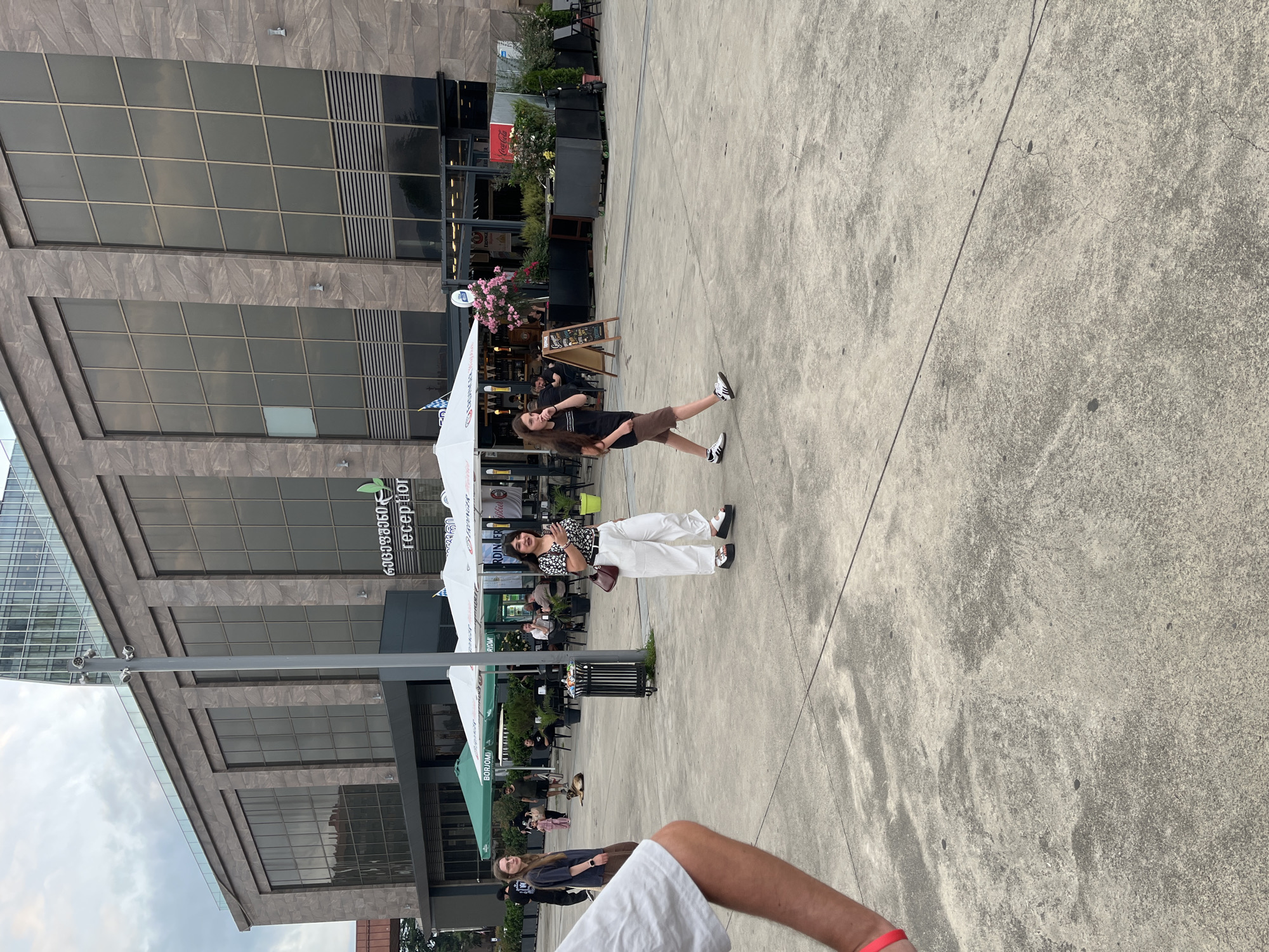 The image depicts an outdoor public space in front of a modern building. A woman in white pants and a patterned top is walking towards the right side of the frame, holding a smartphone. To her left, a younger woman with long dark hair is walking away from the camera, wearing a black t-shirt and brown shorts. In the background, there is a building with large glass windows, some of which reflect the sky. The building appears to be a commercial establishment, possibly a hotel or a restaurant, with signage indicating "reception." There are umbrellas set up in front of the building, suggesting an outdoor seating area. Several people are visible sitting at tables in the background. The ground is paved with concrete. This scene is likely located in Batumi, Georgia, specifically in the area near the Batumi Boulevard or the new port.