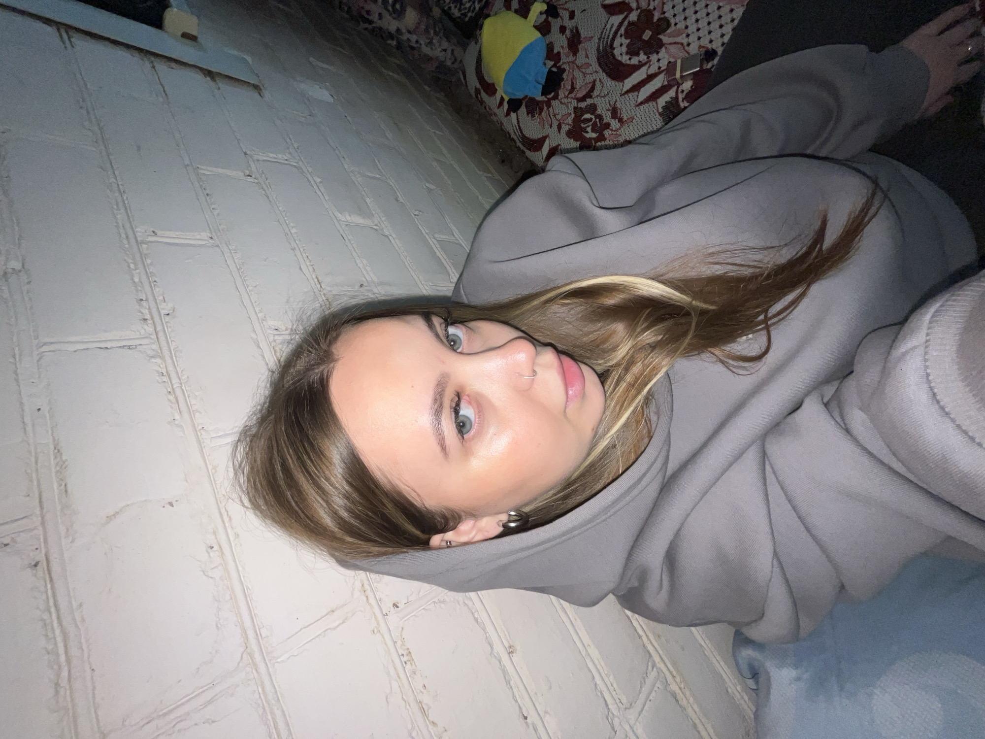 An image, rotated 90 degrees counter-clockwise, shows a young woman lying down in what appears to be an indoor setting in Novopokrovskaya, Russia. She is positioned horizontally, looking directly towards the camera with a neutral expression. Her head is to the left, and her body extends to the right.
The woman has light brown hair with blonde highlights, blue-green eyes, and light skin. She is wearing a grey hooded sweatshirt, with the hood up, framing her face. She has a silver nose ring on her left nostril and multiple earrings in her left ear, including a silver hoop and a larger, sculptural silver earring.
Behind her is a textured wall, possibly painted brick or large rectangular tiles, in a light grey or off-white color. The wall has distinct grout lines or indentations. To her right, a light blue blanket or fabric is visible beneath her arm and body. In the background, partially obscured by her body and the wall, is a patterned fabric, likely a sofa or bed covering, featuring red, white, and brown floral or abstract designs. A small yellow and blue plush toy rests on this patterned fabric in the upper right background. The lighting is dim, suggesting an evening or nighttime setting indoors. There is no visible text in the image.