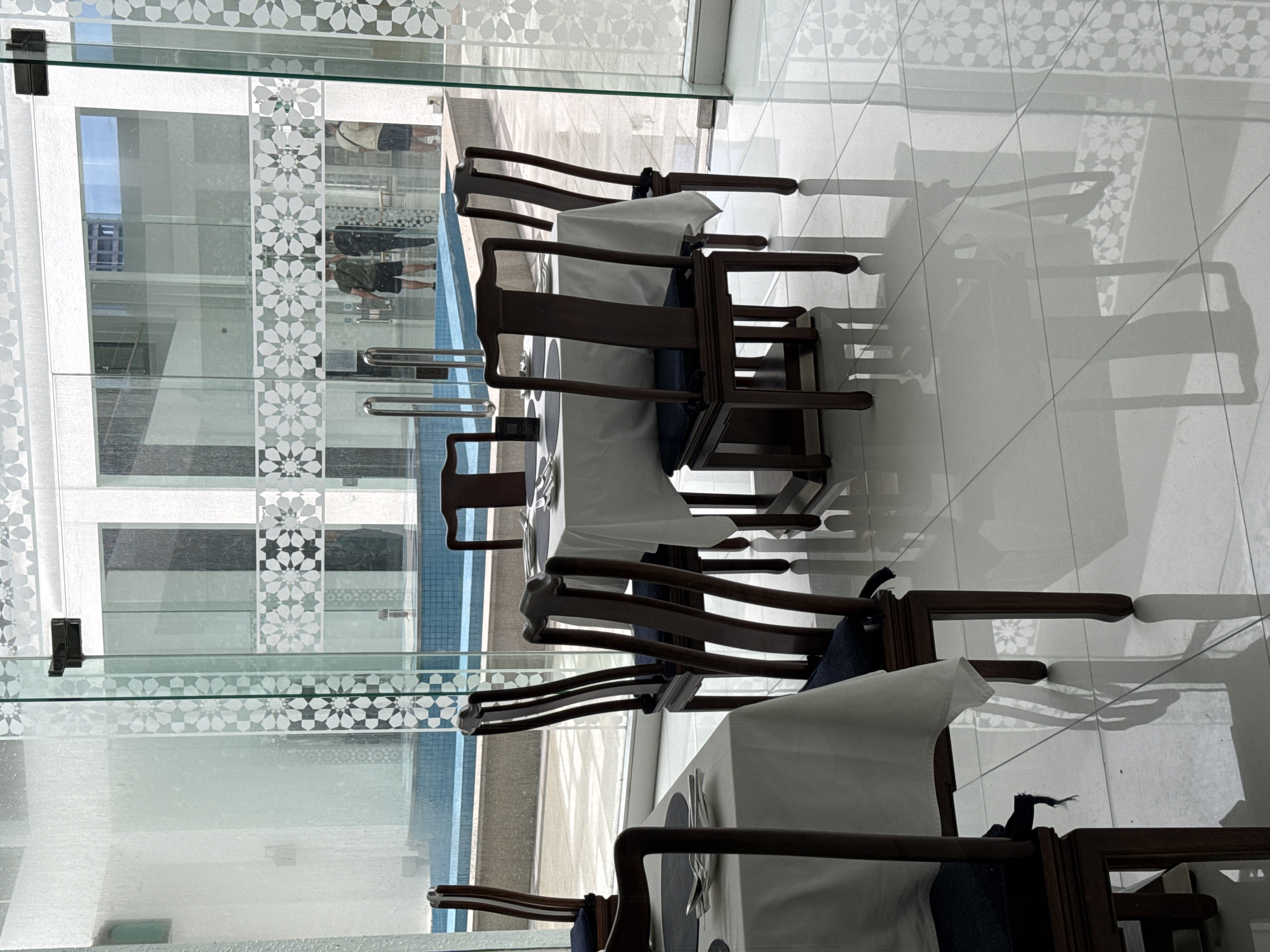 The image shows a restaurant interior with tables set for dining, viewed through a glass wall that reflects a building exterior. The interior features dark wood chairs and tables with white tablecloths. Cutlery is visible on the tables. The glass wall has a decorative pattern of white geometric designs. Reflected in the glass are structures of a building across from the restaurant, and possibly a pool area. Based on architectural styles visible, this scene is likely located in Kuala Lumpur, Malaysia. There are reflections in the glass, appearing to show people walking outside near the pool area, but they are not within the main subject of the image. The focus is on the restaurant tables, chairs, and reflections in the glass.